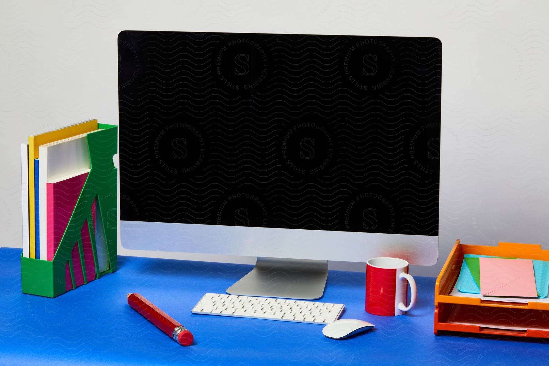 A blue desk with a Mac desktop, keyboard, mouse, coffee, pencil, book holder, and paper shelf in an office.