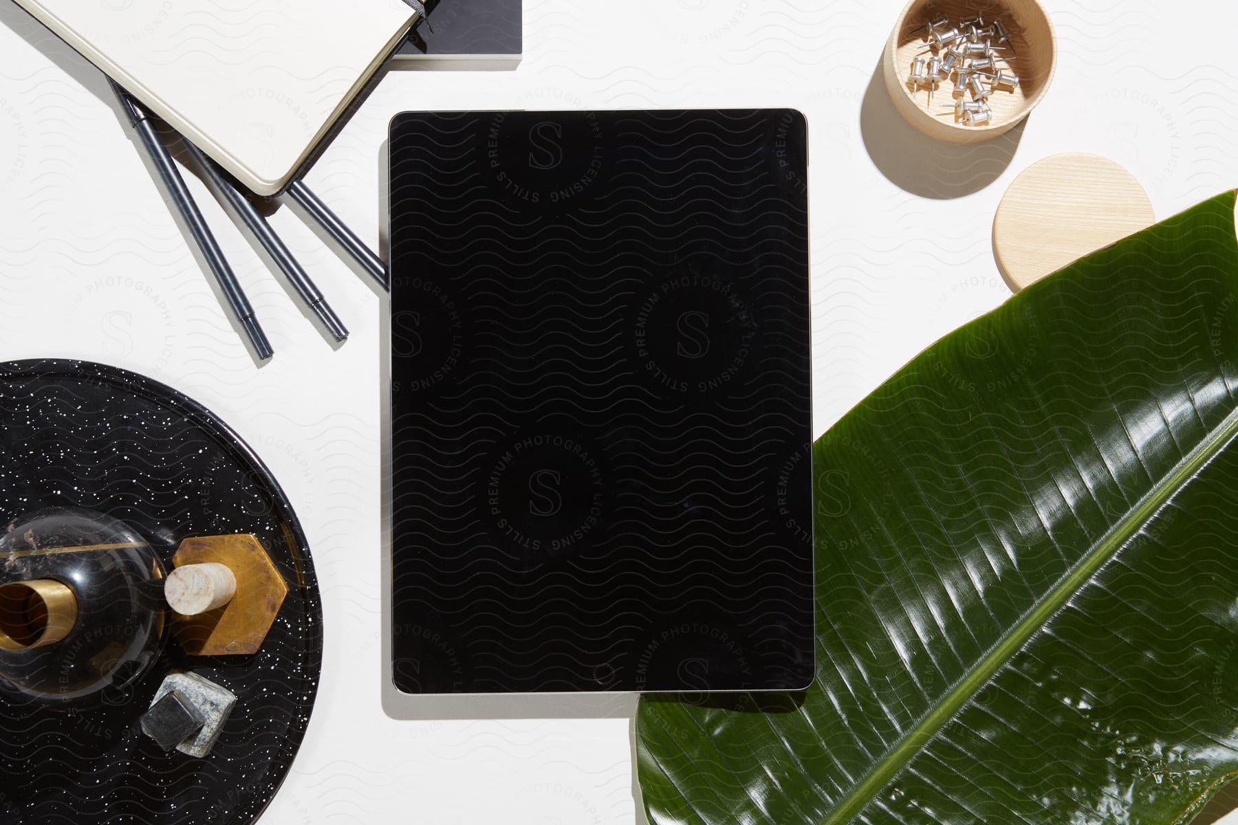 A black tablet on a white counter surrounded by a shiny green leaf and small containers