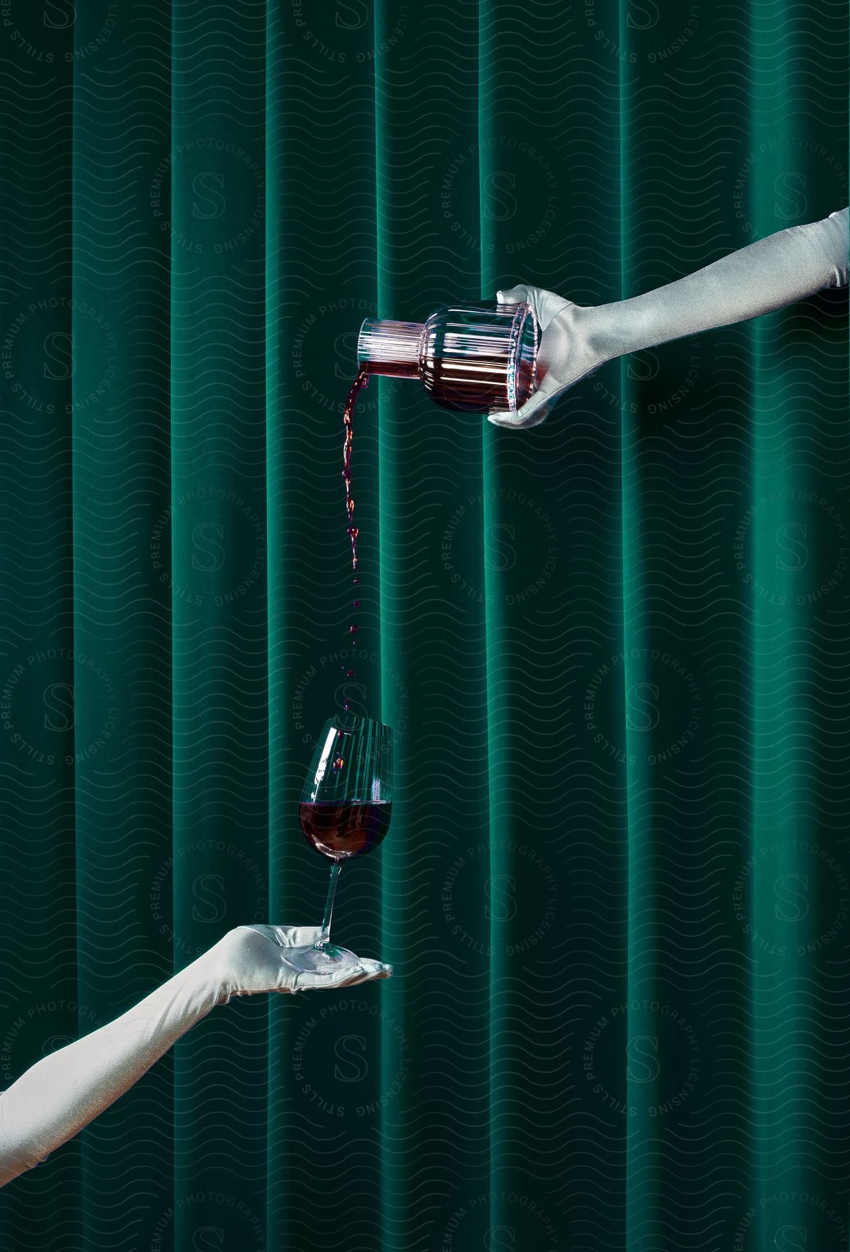 Woman wearing long gloves pours from wine decanter into glass held by another's gloved hand.