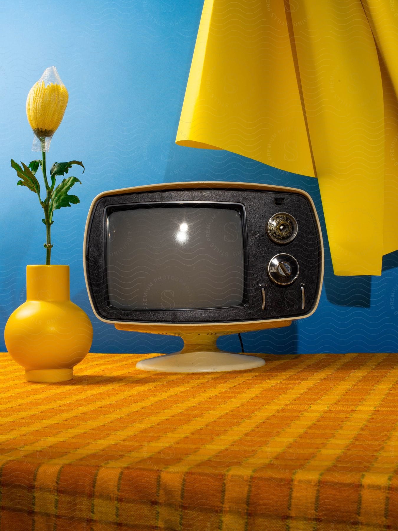 A retro yellow TV sits on a yellow checkered table with a yellow vase and flower, against a blue wall.