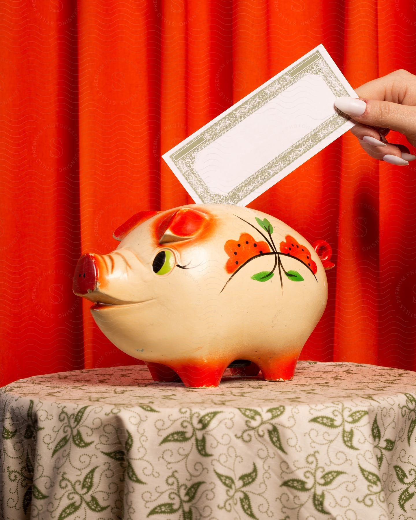 A woman's hand deftly slides some money into a piggy bank, which sits proudly on a wooden table in front of red curtains