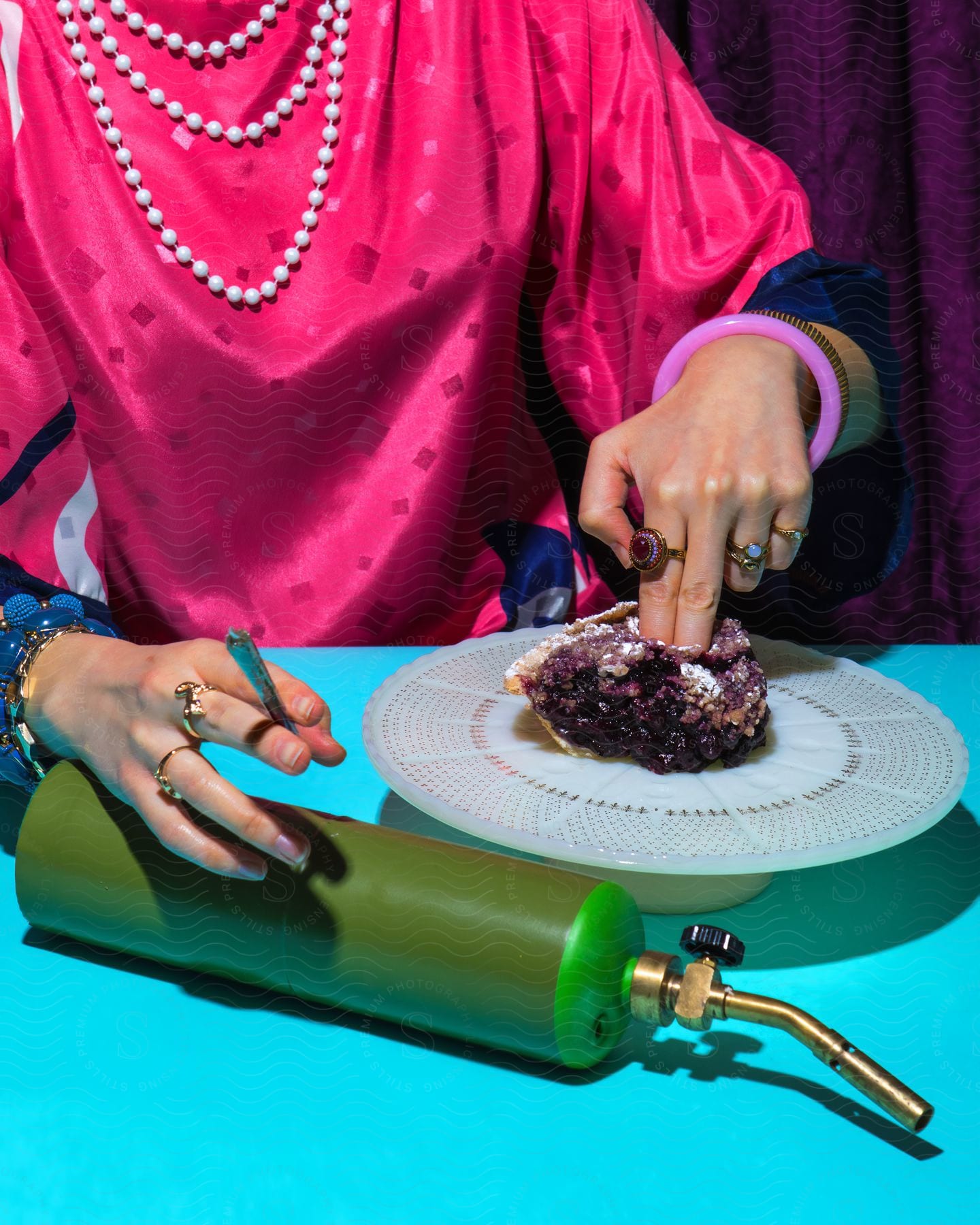 A woman in a flowing pink garment presses her fingers into a piece of blueberry pie on a table next to a blowtorch.