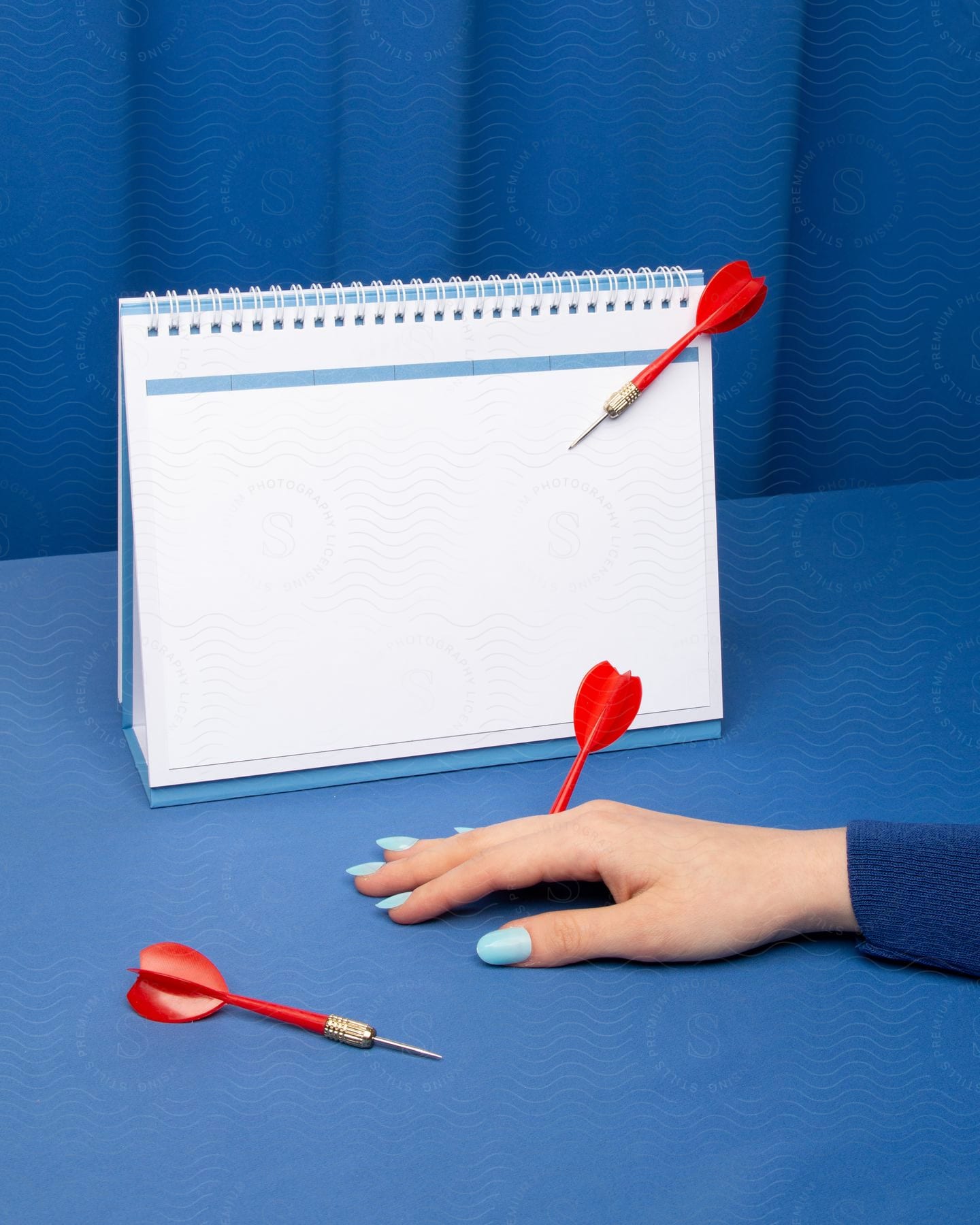 A woman poses her hand with painted nails while red darts are placed around a calendar.