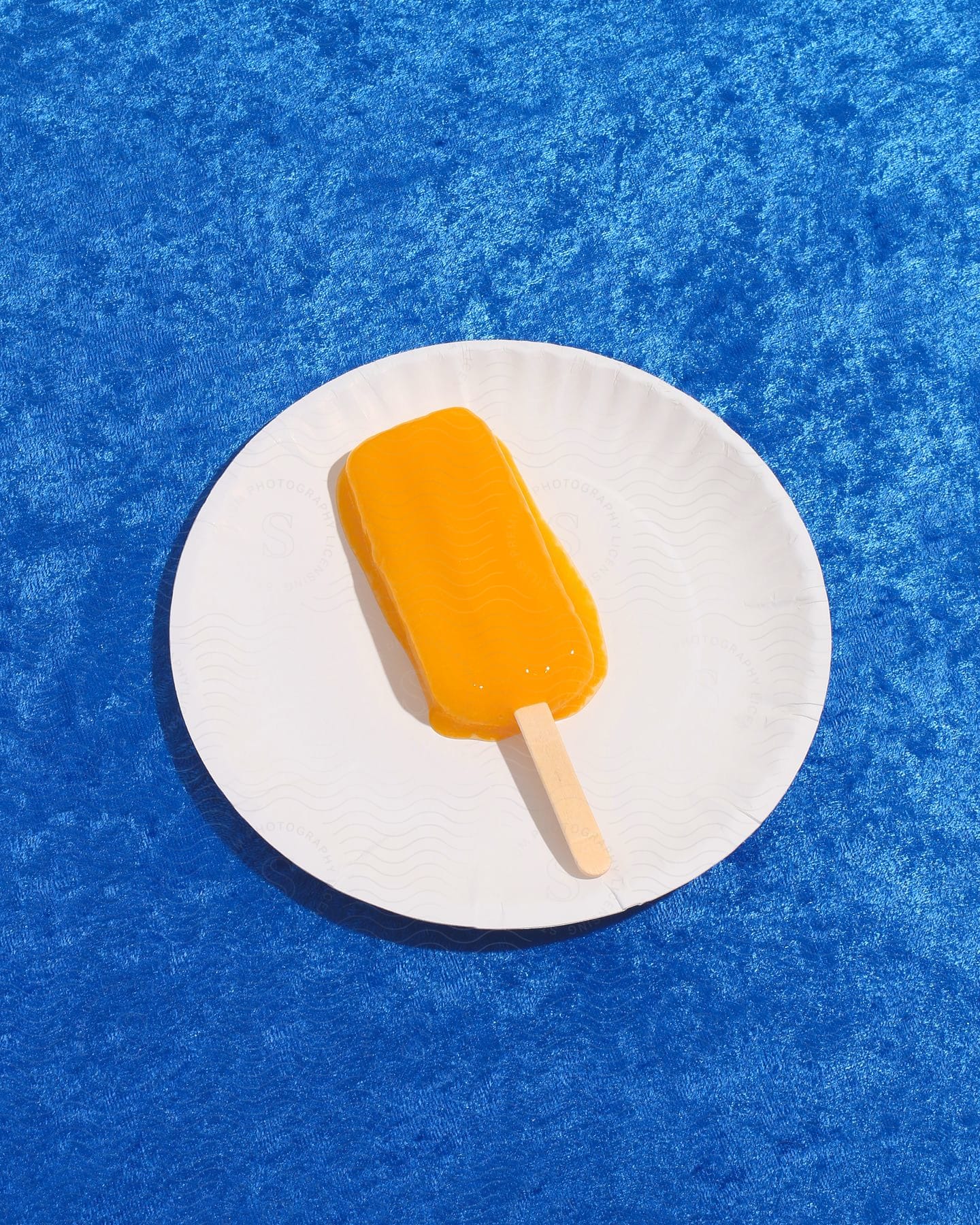 Stock photo of a melting orange popsicle on a paper plate on a blue surface.