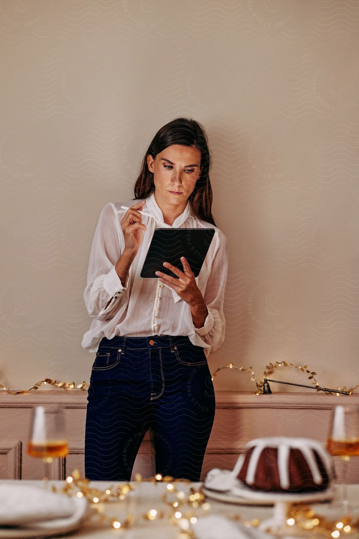 Stock photo of woman concentrates on tablet while standing near table holding glasses and a cake.