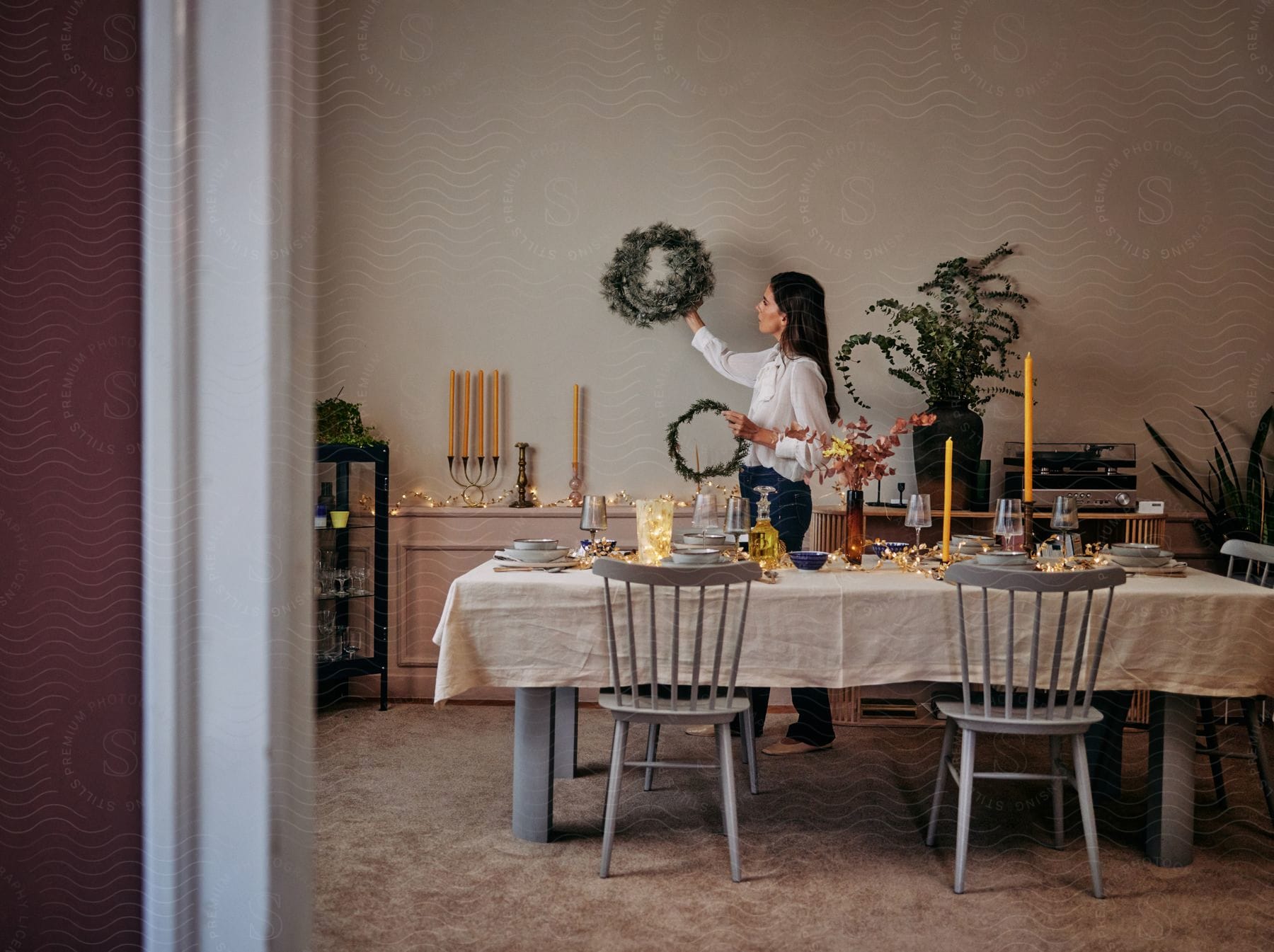 A dining room table is set for a party as a woman puts a wreath on the wall