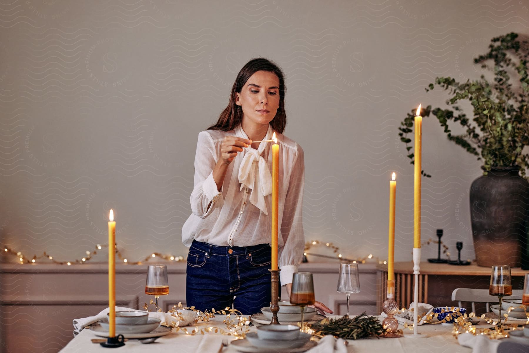 A woman wearing a white blouse and blue jeans stands at a table set with candles, plates, cups, food, and beverages, using a match to light a tall orange candle.