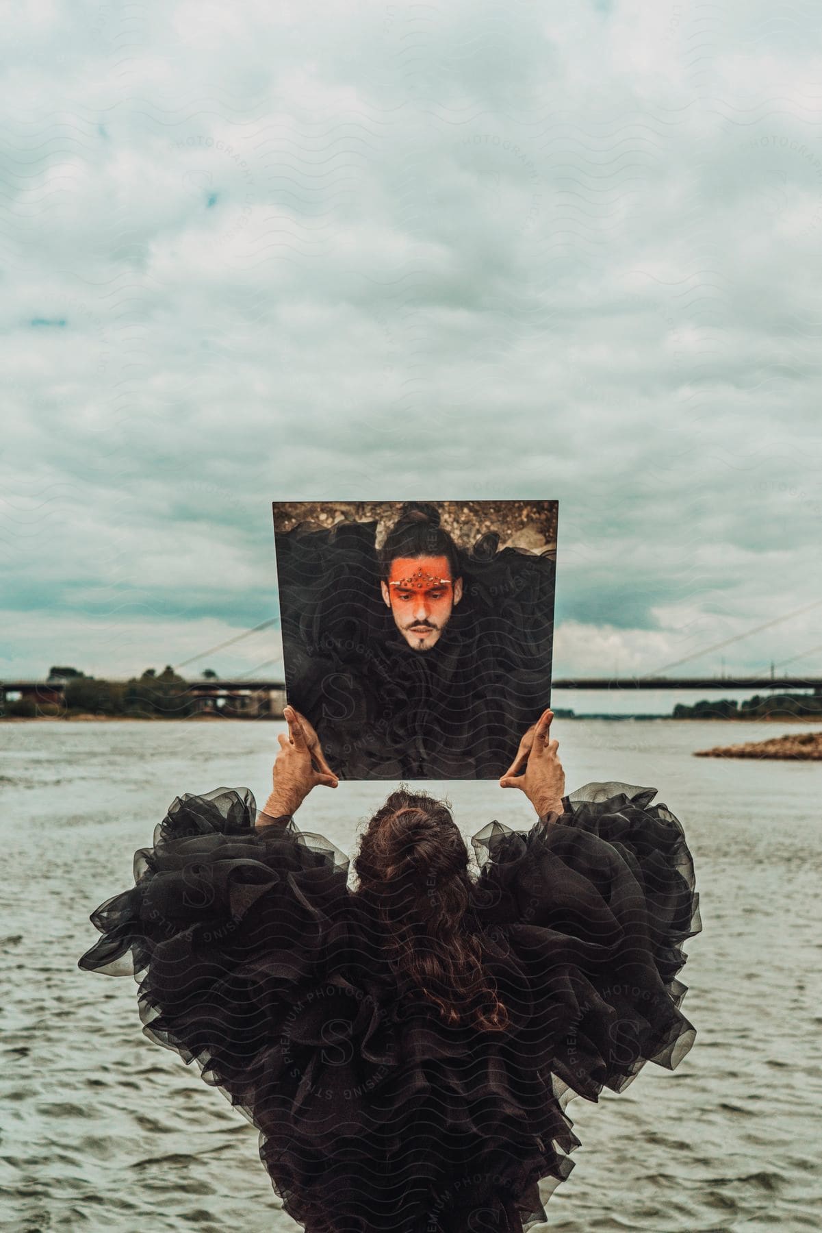 Man standing on shore holds up mirror to reflect elaborate makeup and attire.