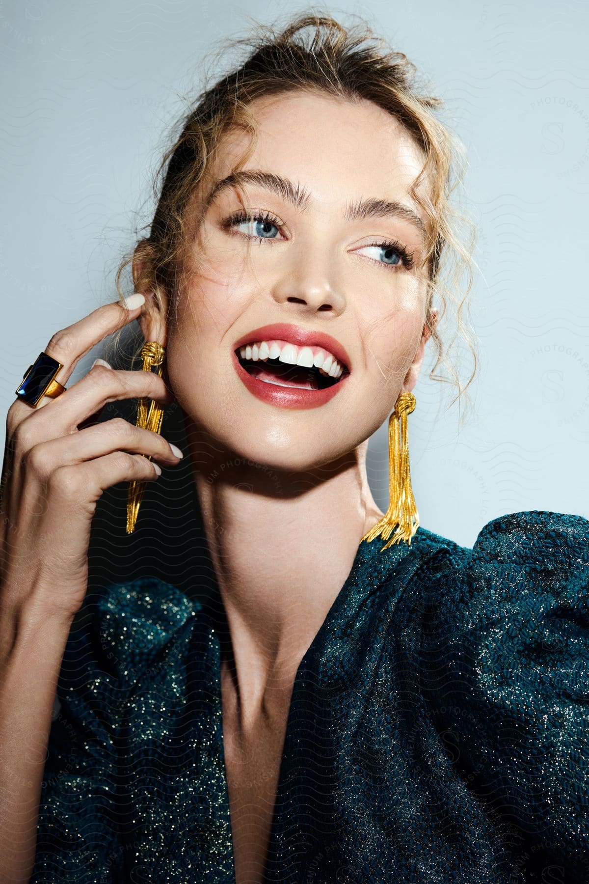 A female model wearing a dark dress and long earrings smiles while holding her hand to her face.