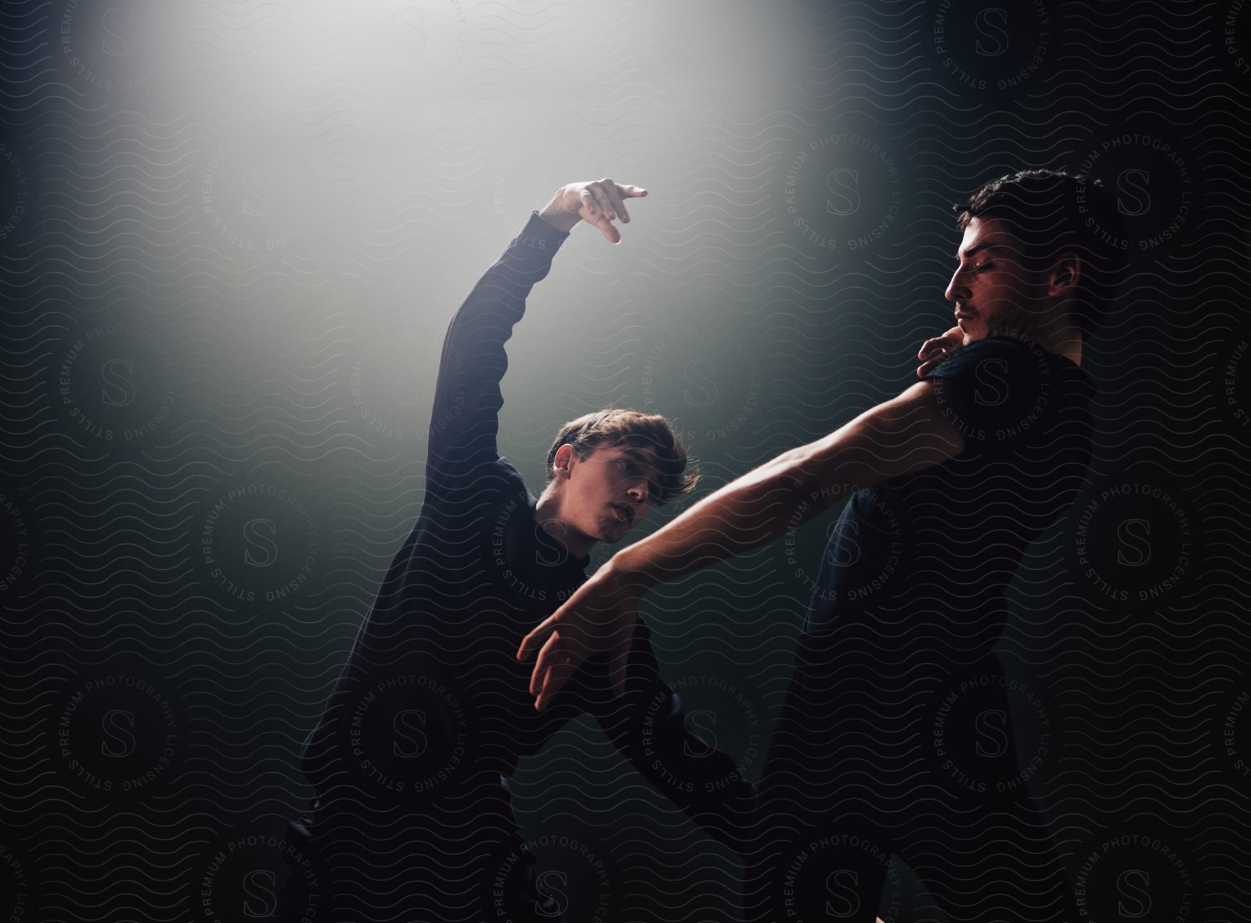 Two men wearing shirts dance in a spotlight in a dim-lit room.