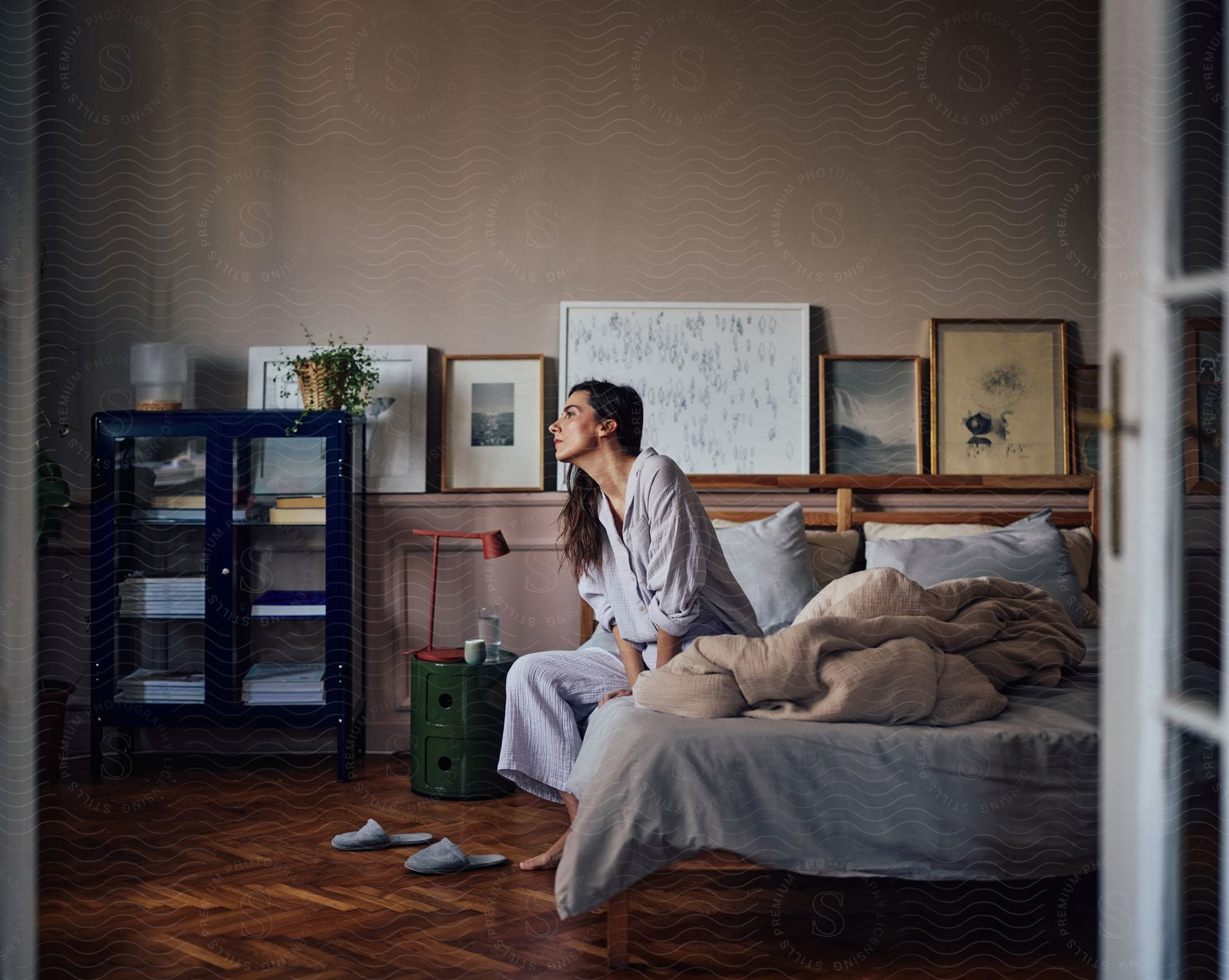 Woman in light blue pajamas sits on unmade bed with a wooden headboard.