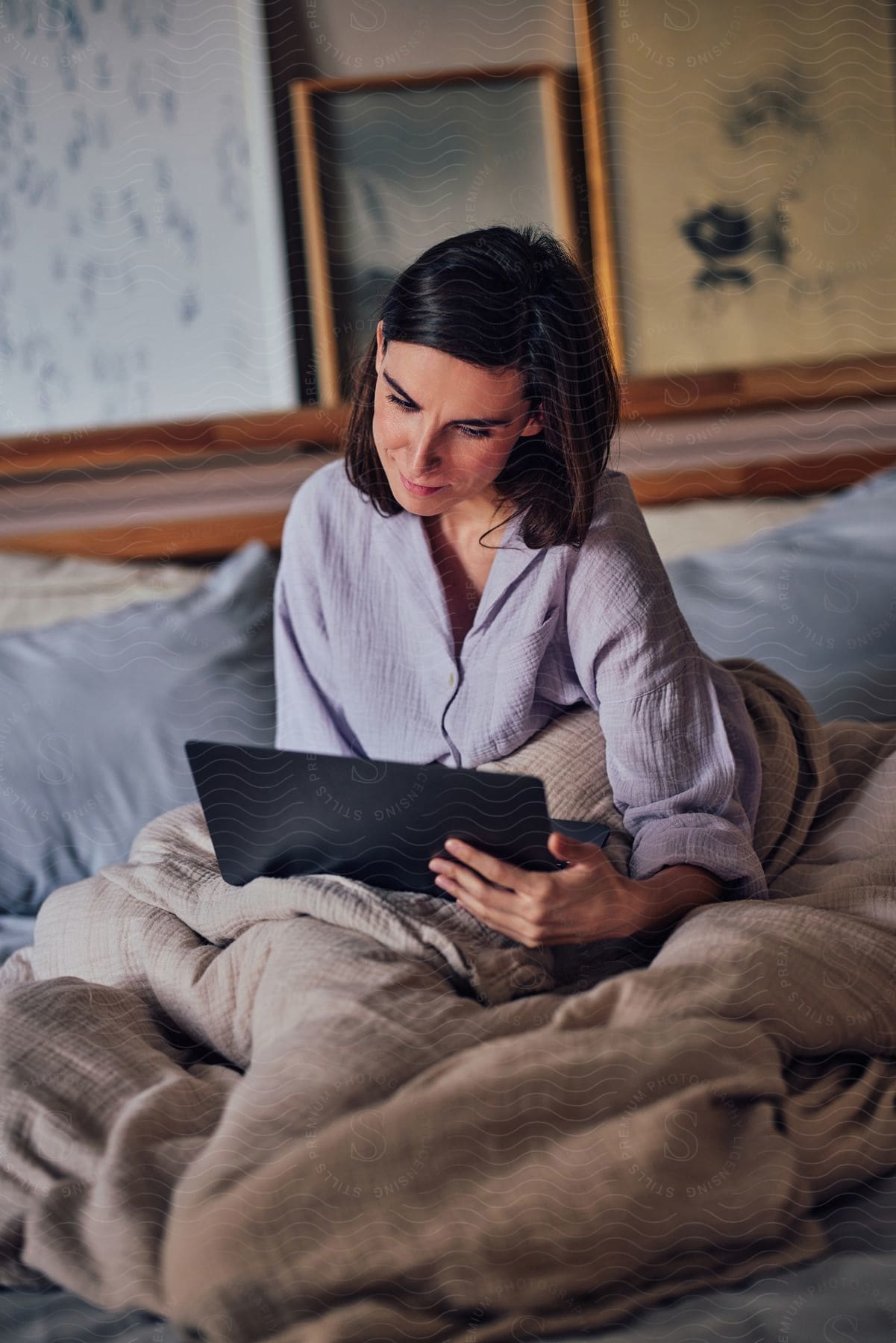 A woman focused on a laptop screen, sitting up in bed with bedding pulled up to her waist.