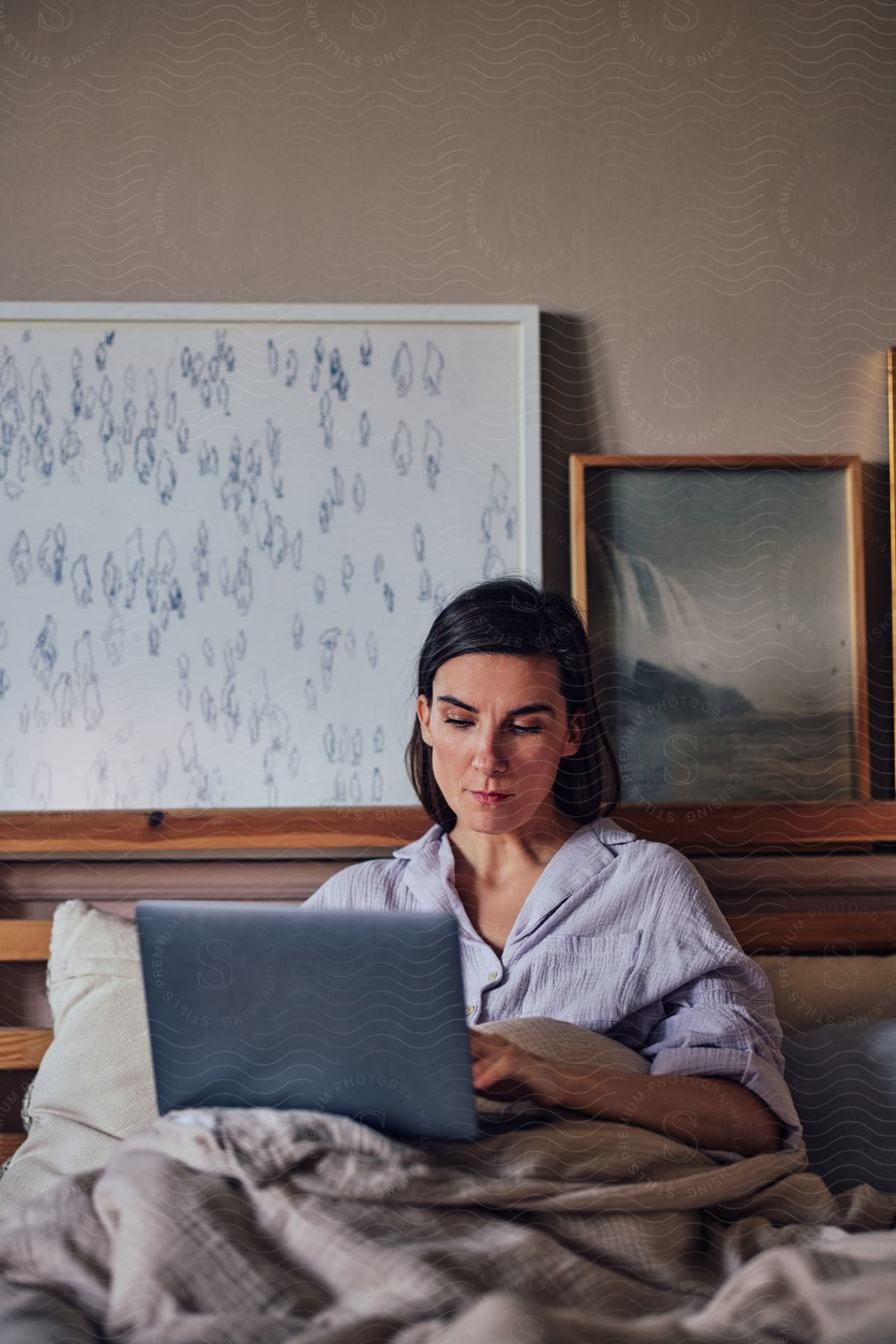 A woman focused on a laptop screen, sitting up in bed with bedding pulled up to her waist.