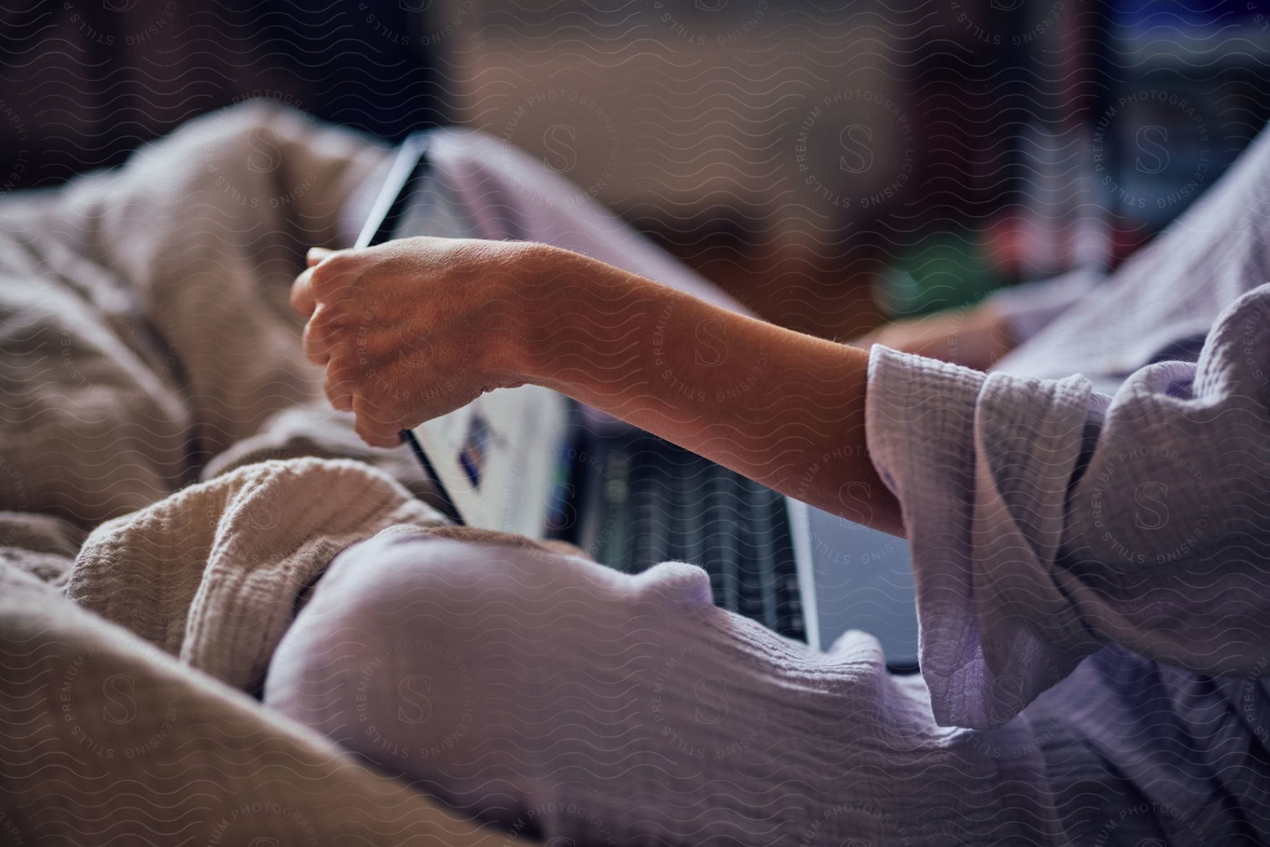 Woman in pajamas using a laptop while sitting in bed, focus on forearm and hand.