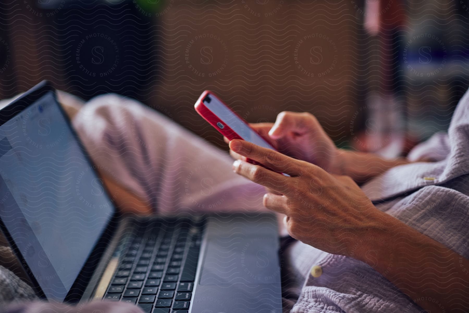 Woman with light blue pajamas sits in bed using a smartphone and a laptop in bedroom