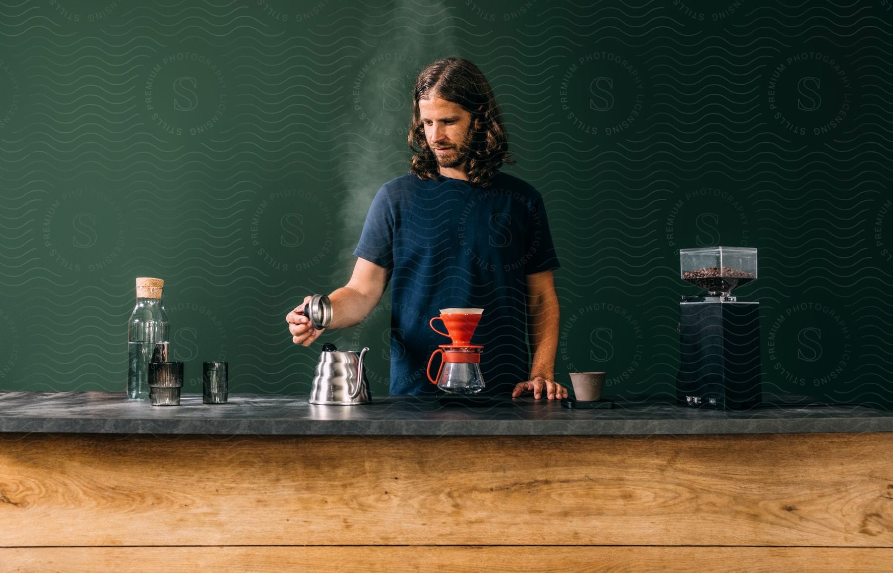 A man is standing behind a counter with containers and a coffee pot as he holds the lid to a teapot and steam rises