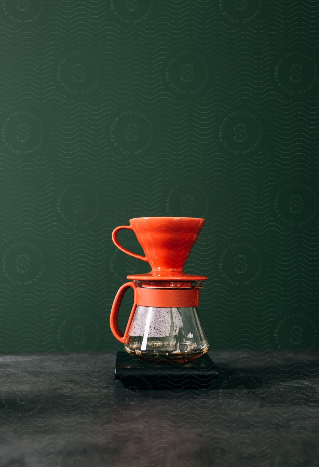 A tapered, orange coffee mug rests on top of a coffee pot on a grey countertop.