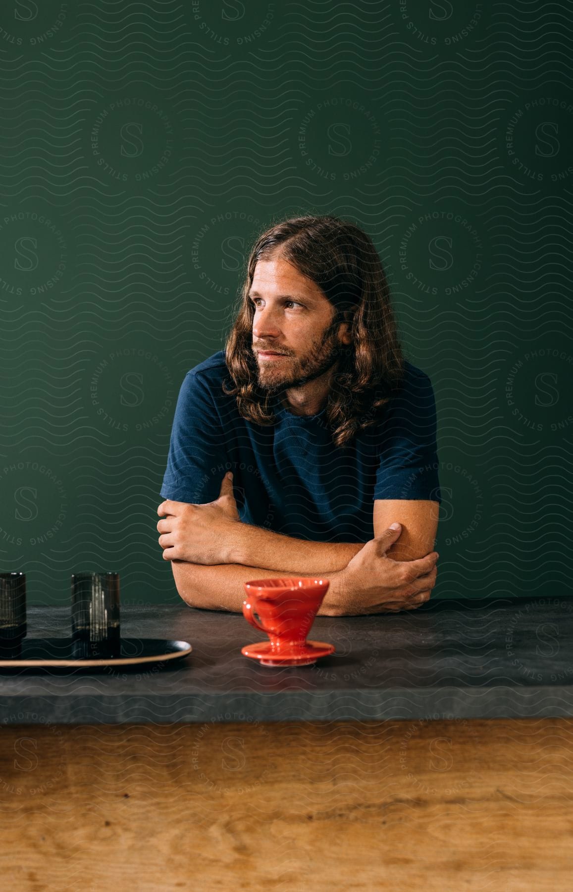 A long haired man with a beard gazes to his right while drinking coffee from a red cup and leaning on the countertop.