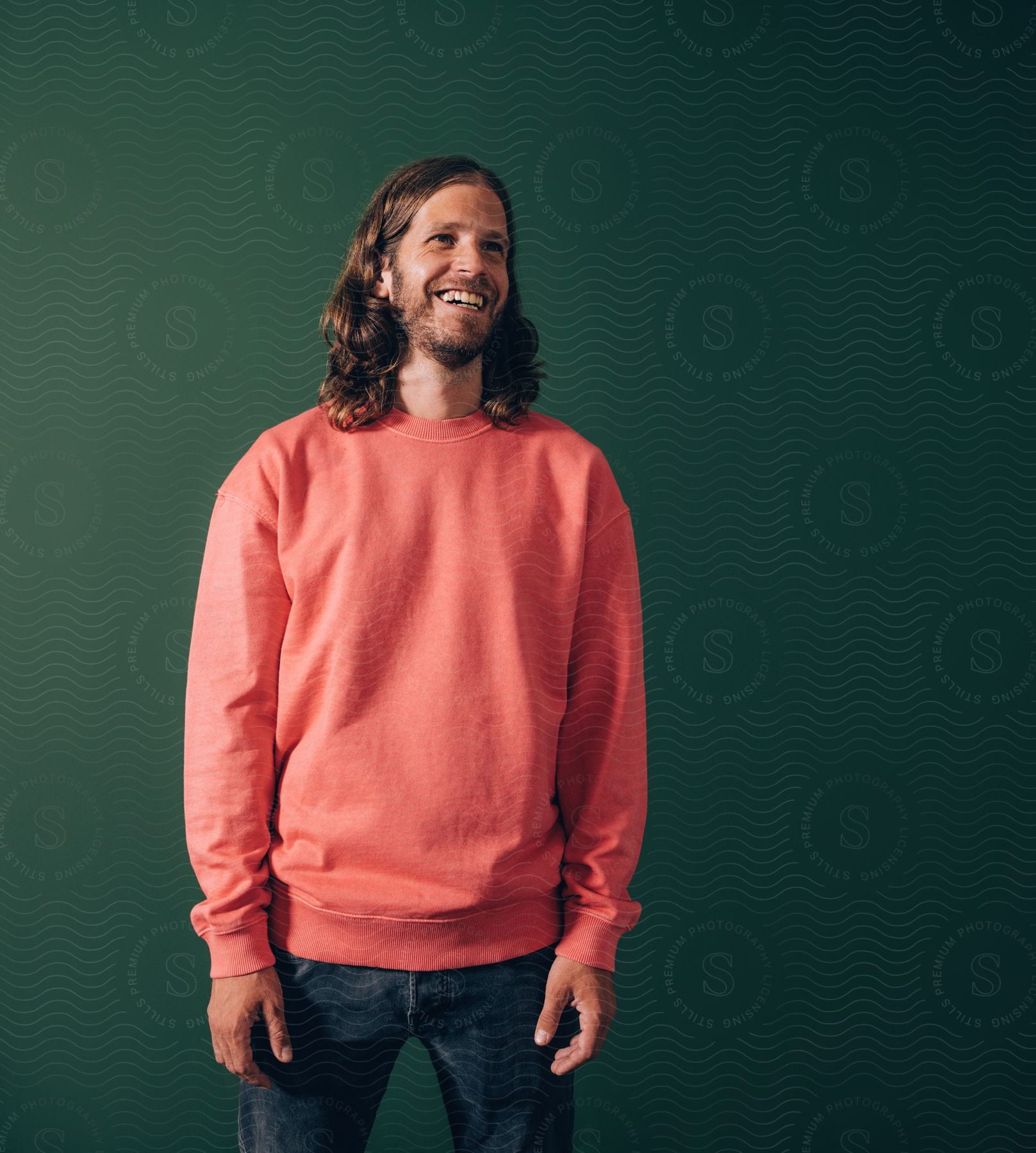 A bearded man with long hair a red sweatshirt and jeans stands in front of a green wall