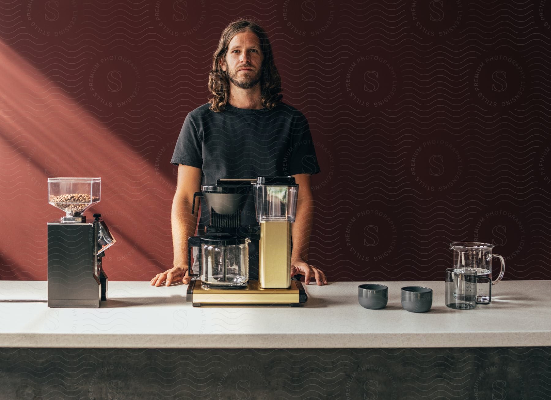 A man with long hair looking straight ahead and below him there is a table with some coffee making machines.