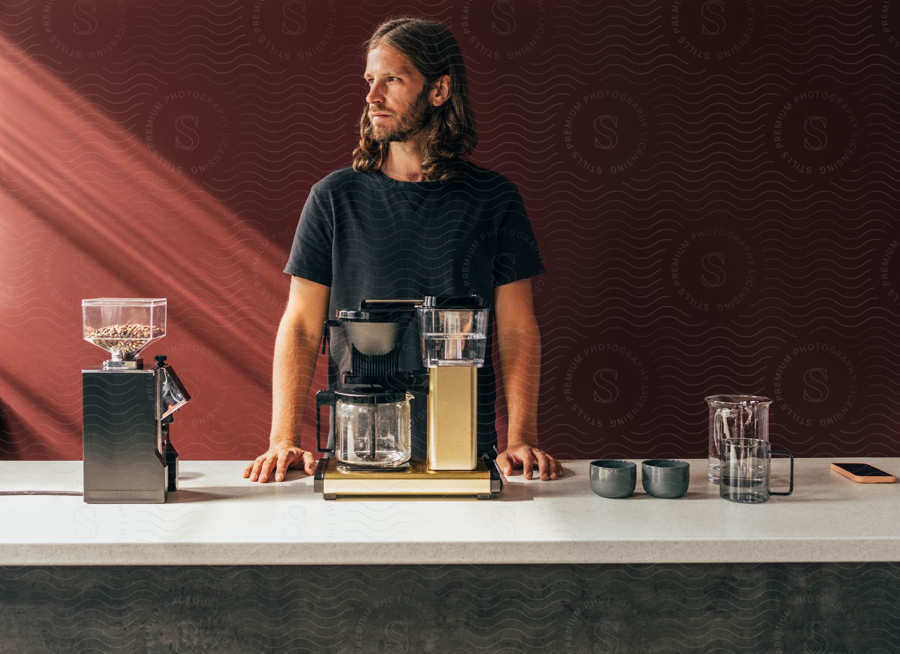 A bearded man with long hair, dressed in a black t-shirt, stands behind a counter with a coffee machine.