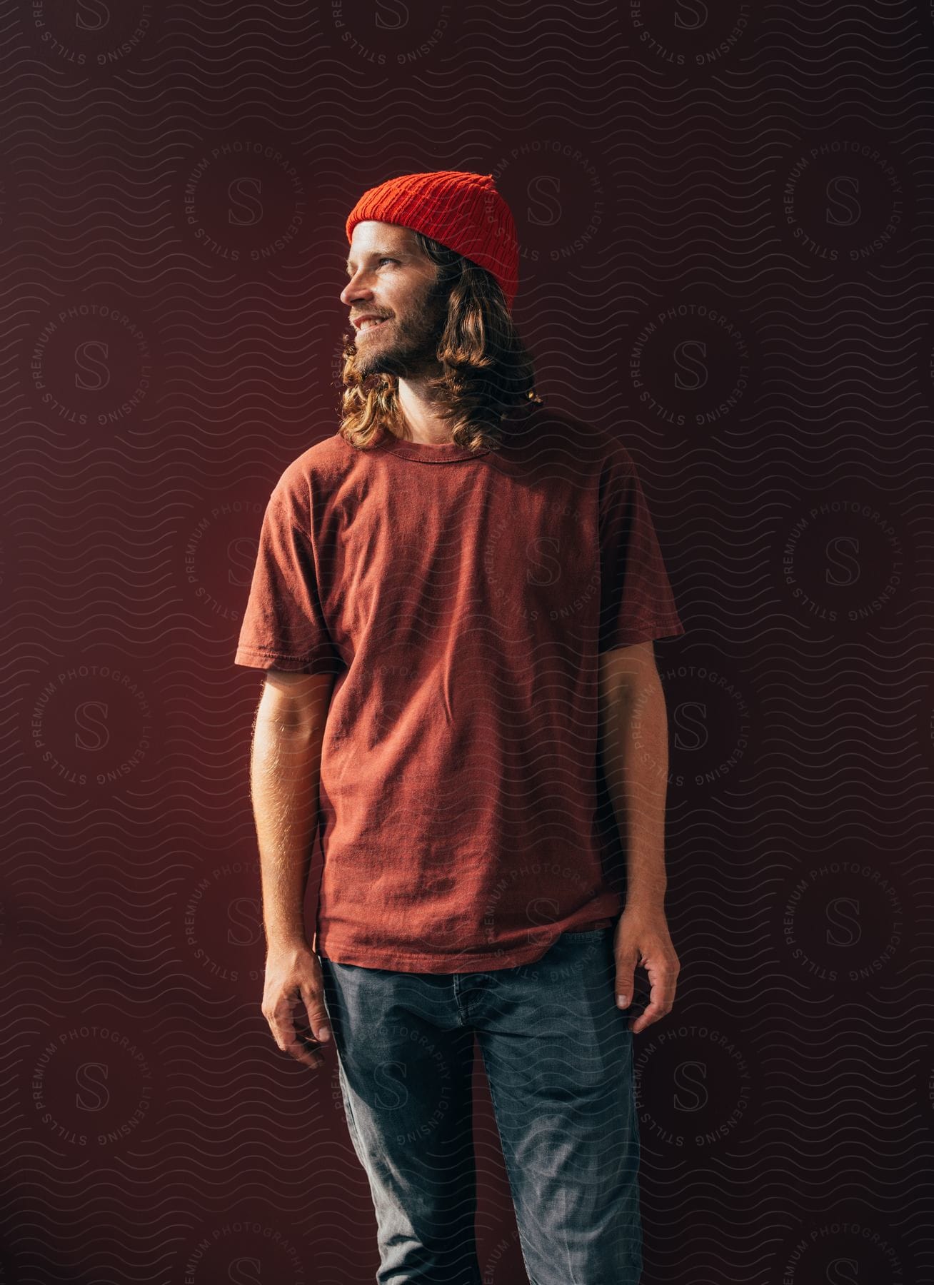 young adult with a red head warmer and a brown shirt stands against a brown wall