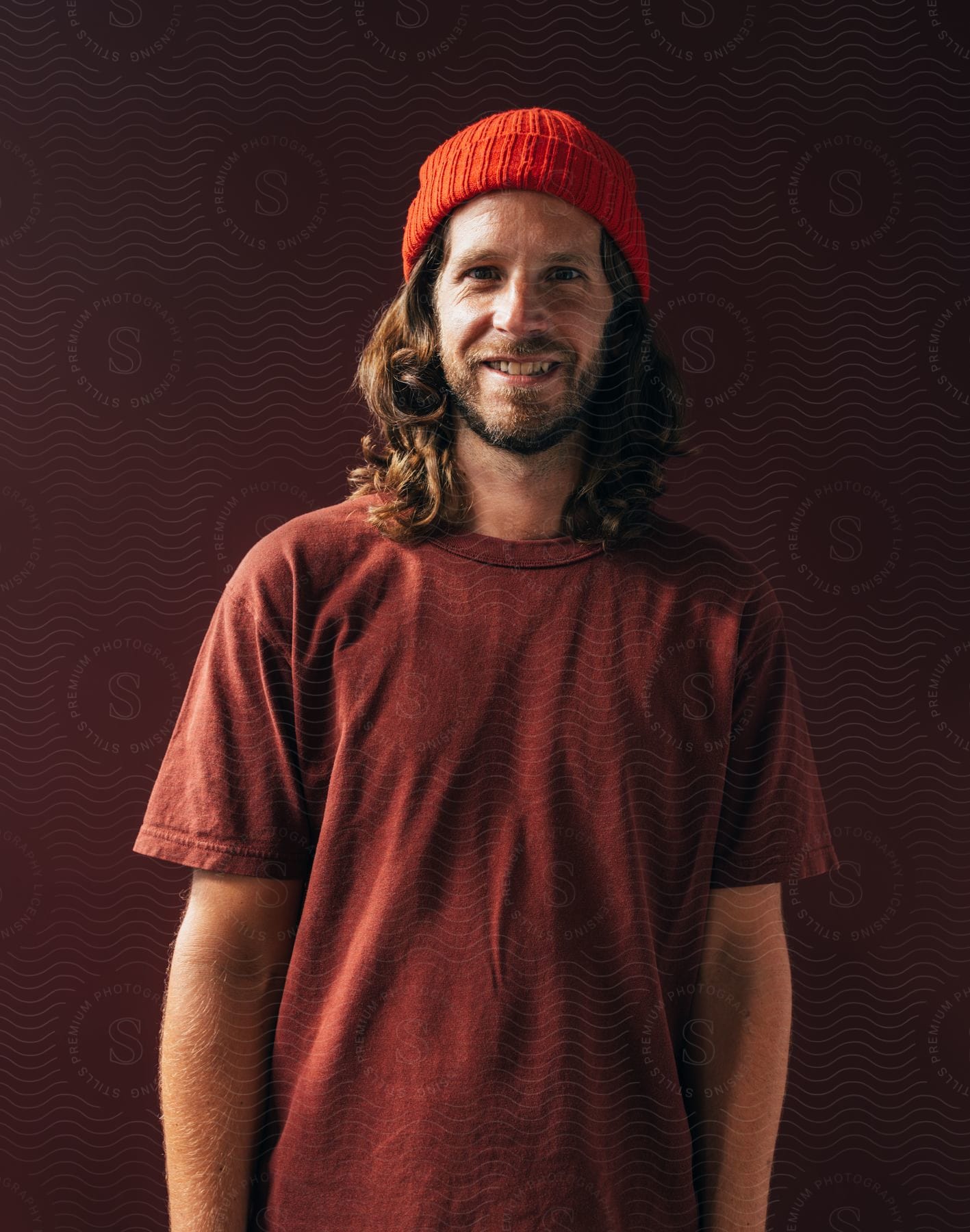 A man with shoulder length hair wearing a t-shirt and beanie stands smiling