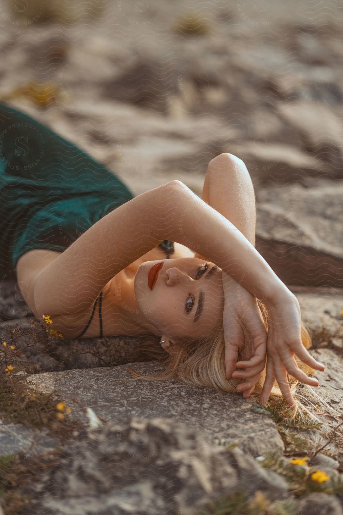 A blonde model in a green gown and makeup posed on her back on stones in the sunlight