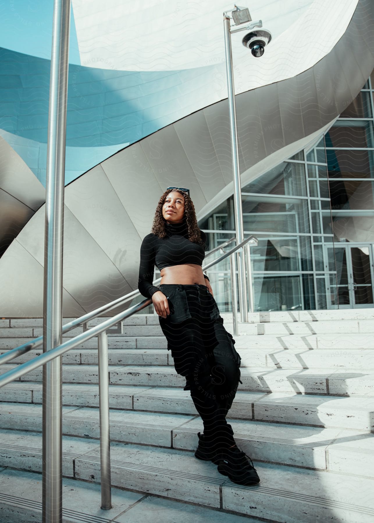 A woman wearing black pants and a long sleeved halter top is standing on steps outside of a building leaning against the handrail