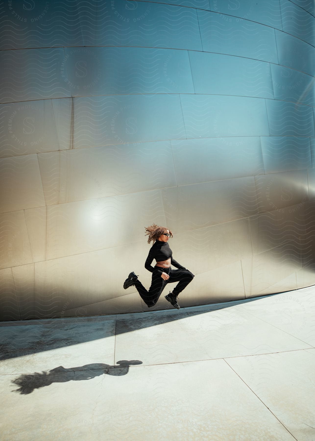 A women is  jumping in the air in front of a concrete wall with her legs in a running motion.