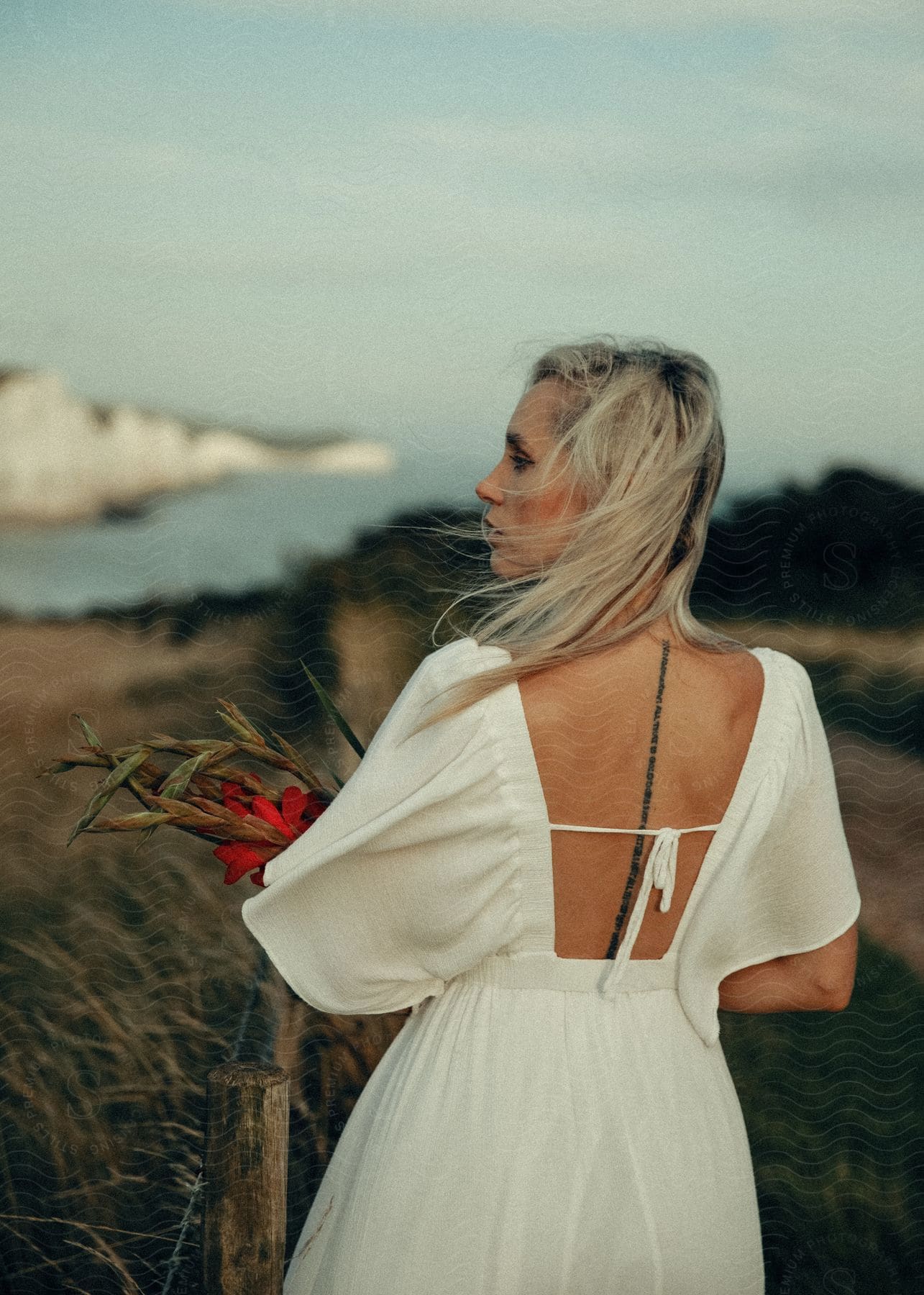 A blonde woman wearing a white dress and holding red flowers stands against the coast.