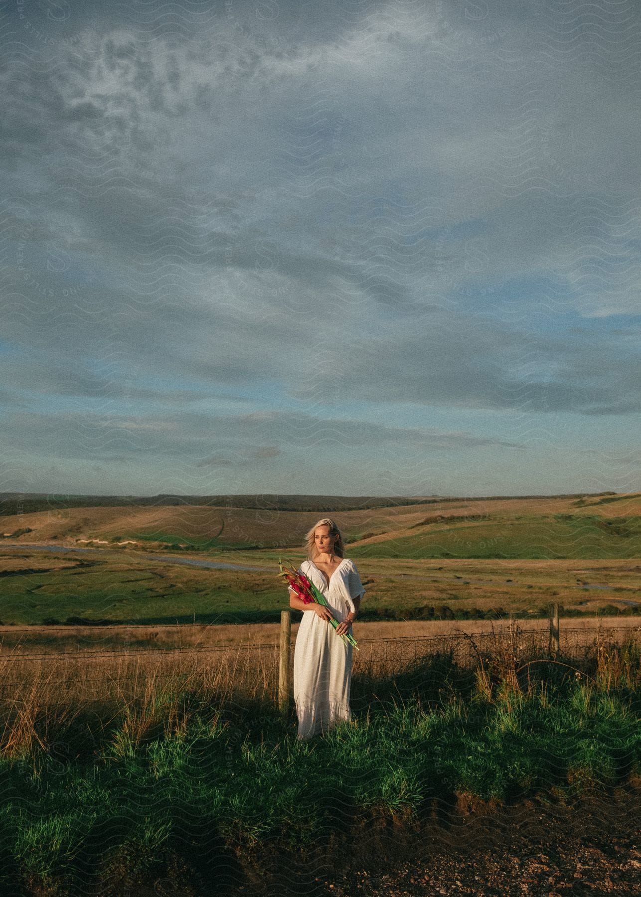 A woman with blonde hair wearing a long white dress is standing on rural land holding a bouquet of flowers