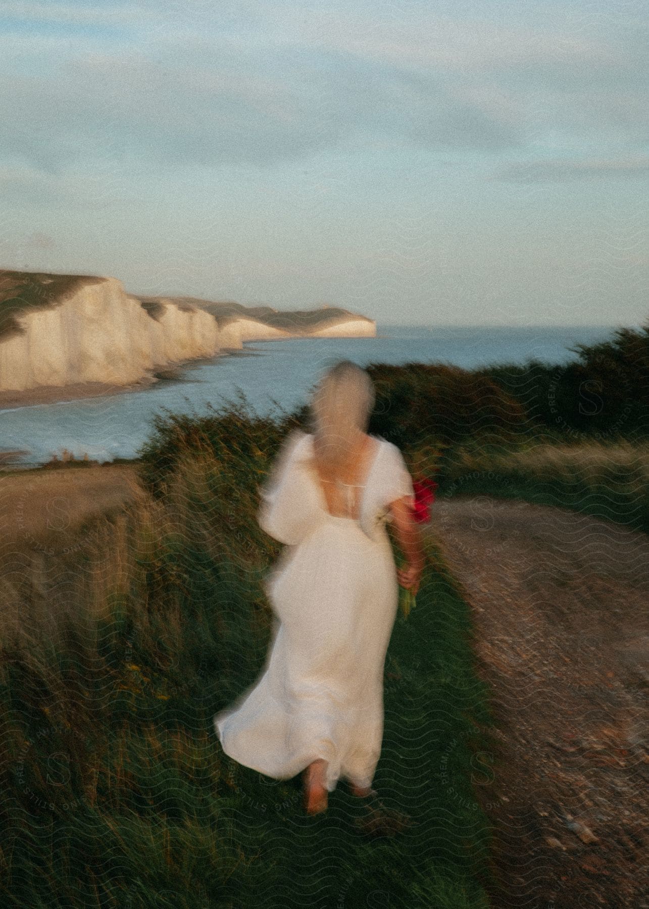 Blurry fashionably gowned figure of woman holding bouquet walks along road across water from cliff.