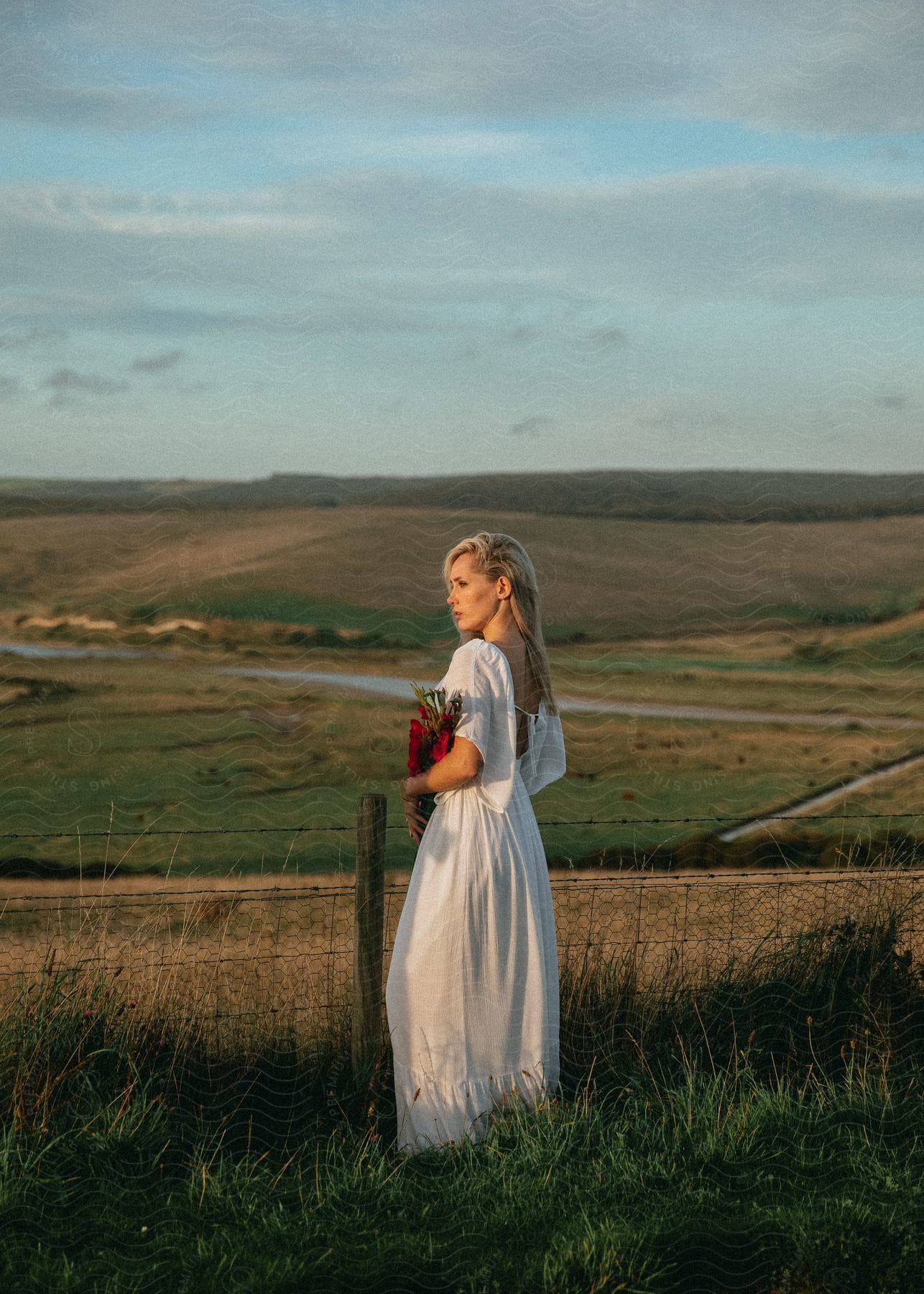 Blonde woman in a white dress holding a bouquet of flowers in an open field.