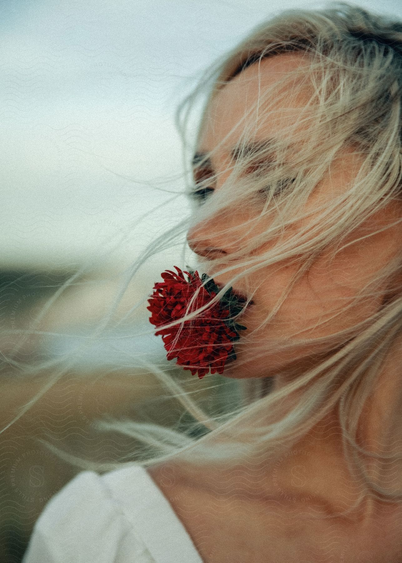 A woman stands outside with a red flower in her mouth as her long blonde hair blows across her face