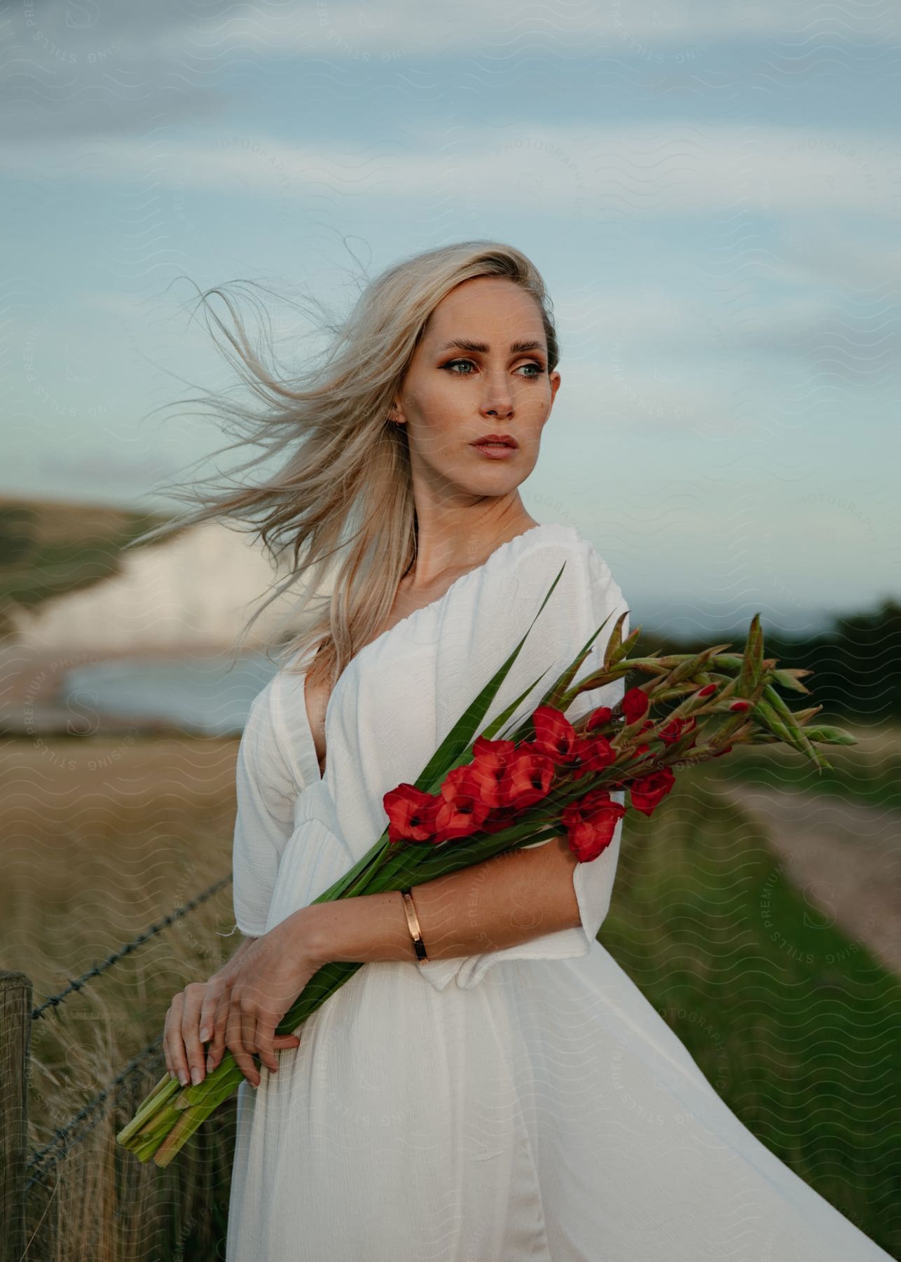 A woman with blonde hair wearing a white dress is standing near the coast holding a bouquet of flowers