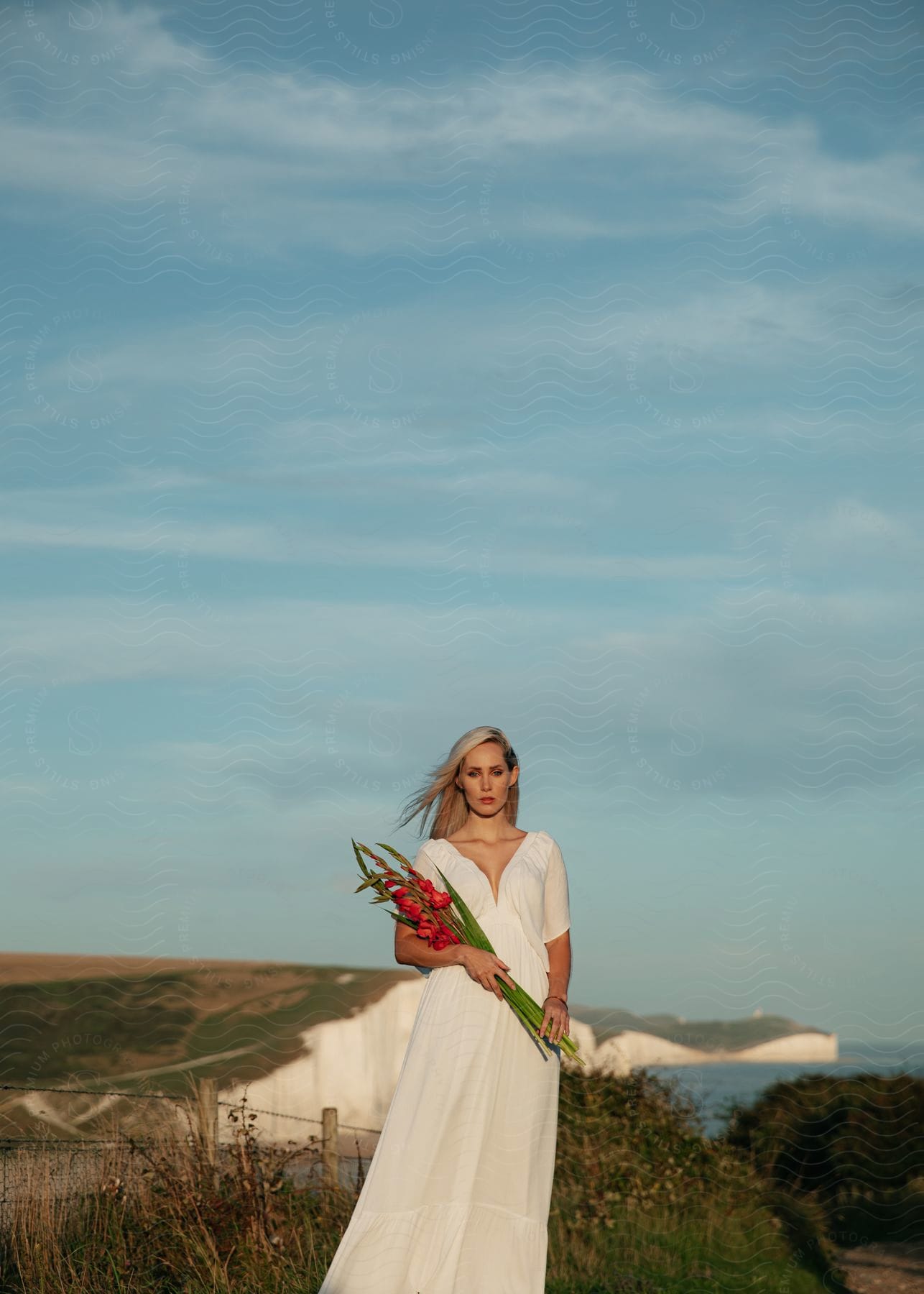 A woman with blonde hair wearing a long white dress is standing along the coast holding a bouquet of flowers