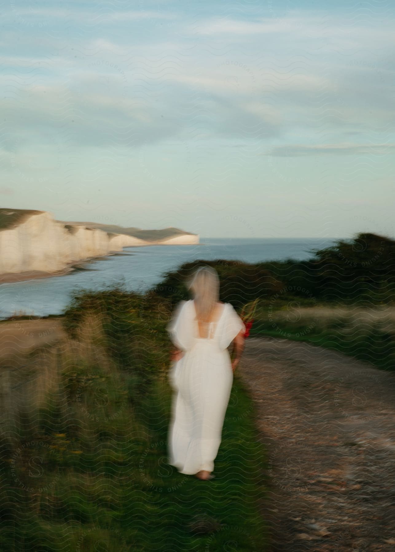 A woman with blonde hair wearing a long white dress is walking next to a dirt road along the coast