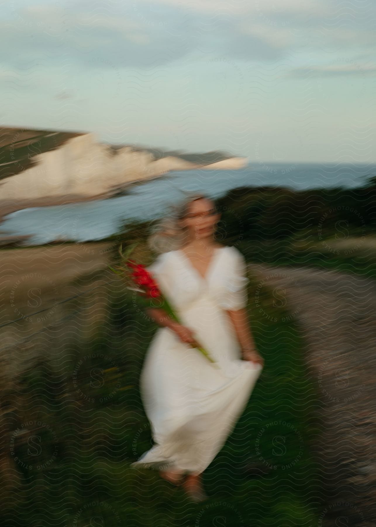 A woman with blonde hair wearing a long white dress is sitting along the coast holding a bouquet of flowers