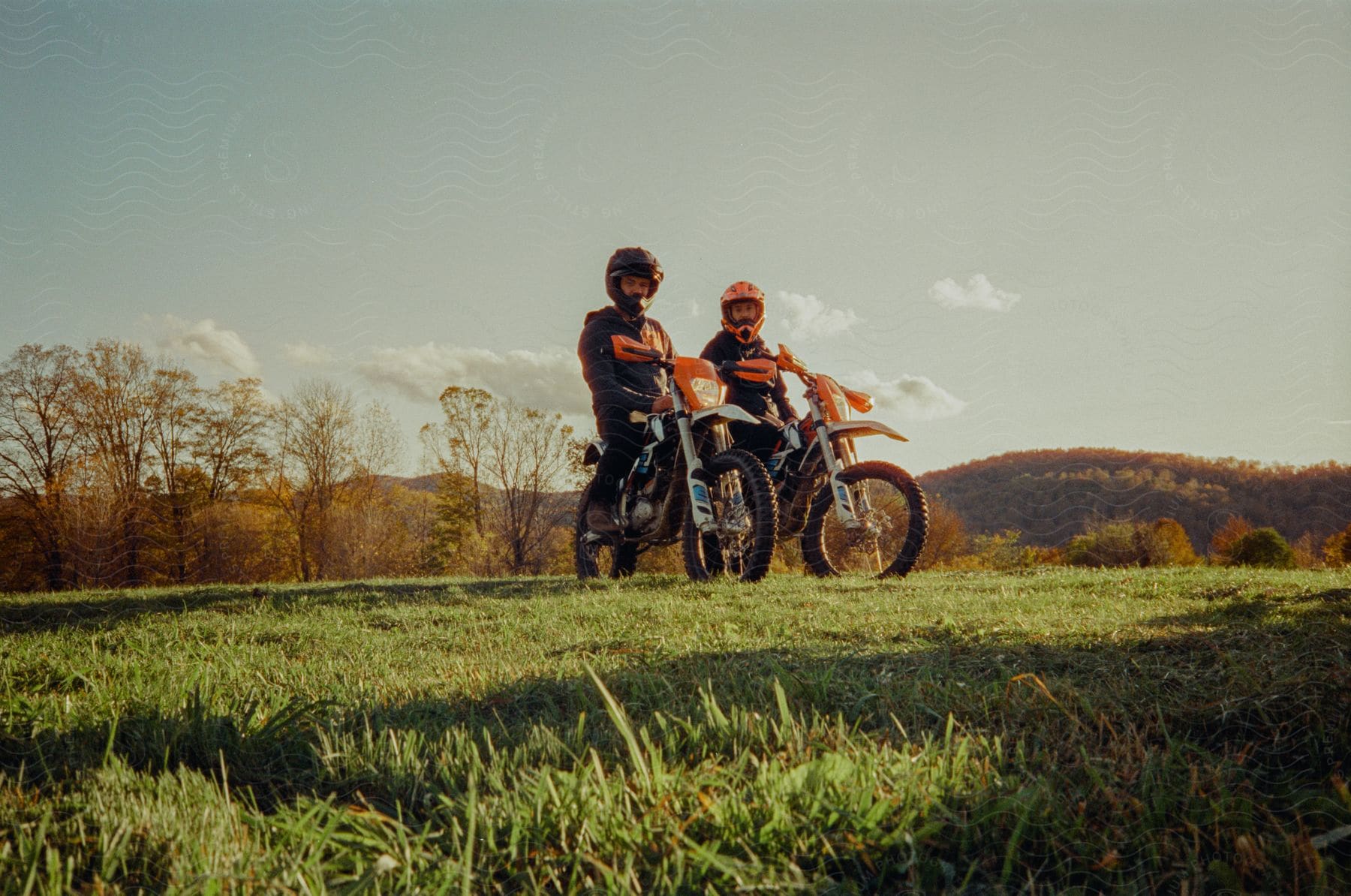 Men wearing helmets and jackets sitting on dirt bikes in a green field.