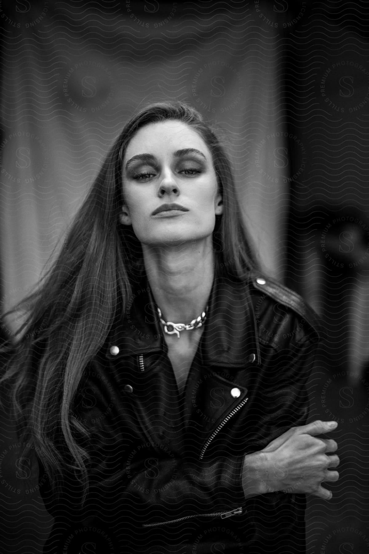 Woman in a leather jacket with a chain necklace poses against a draped fabric background.