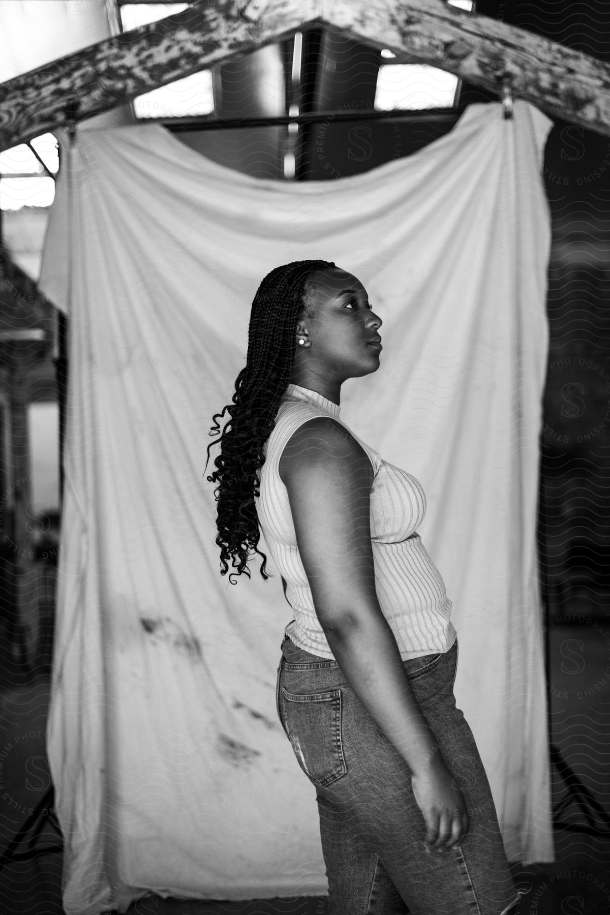 A woman with curly hair, wearing a white top and jeans, stands in front of a dirty curtain in a room