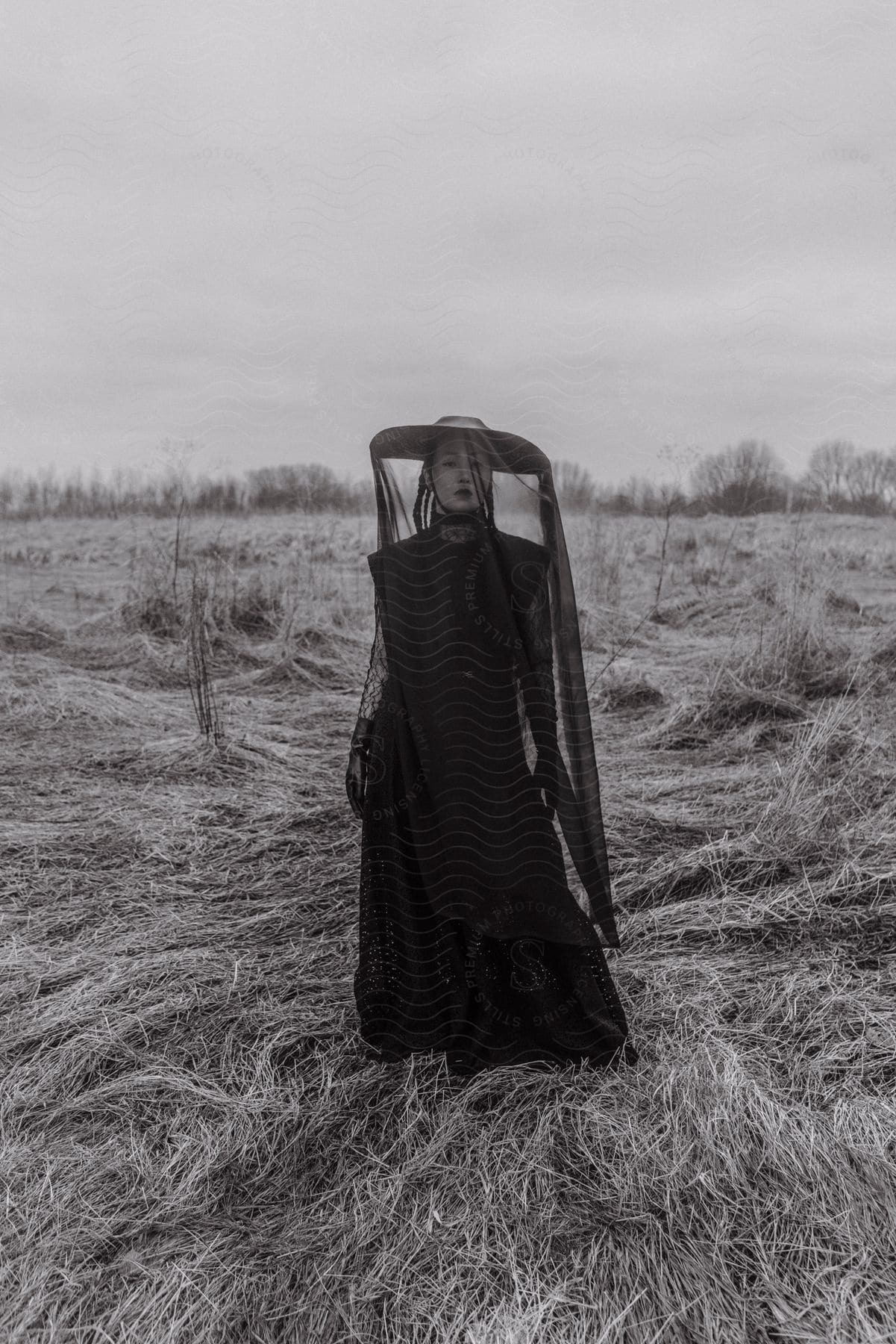 A woman is standing in a field wearing a long black dress with gloves and a hat with a black veil over the brim