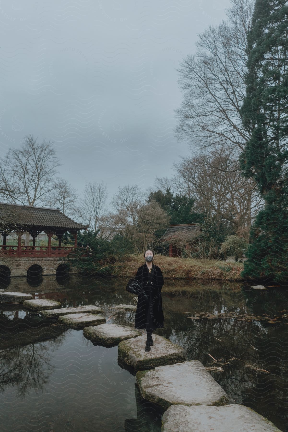 A woman wearing a long black coat and a face mask is walking on rocks across a river