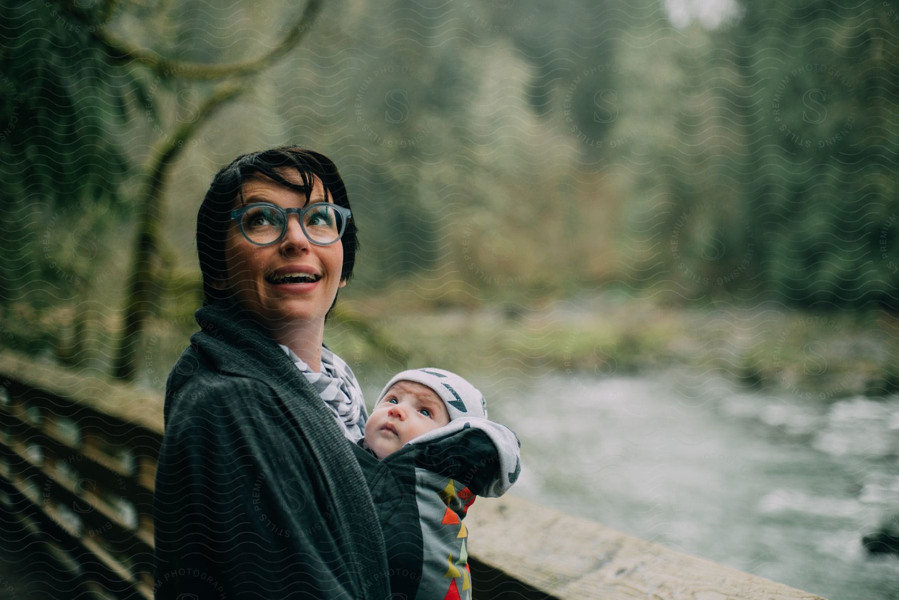 A women is baby wearing an infant boy and standing on a bridge over a creek.