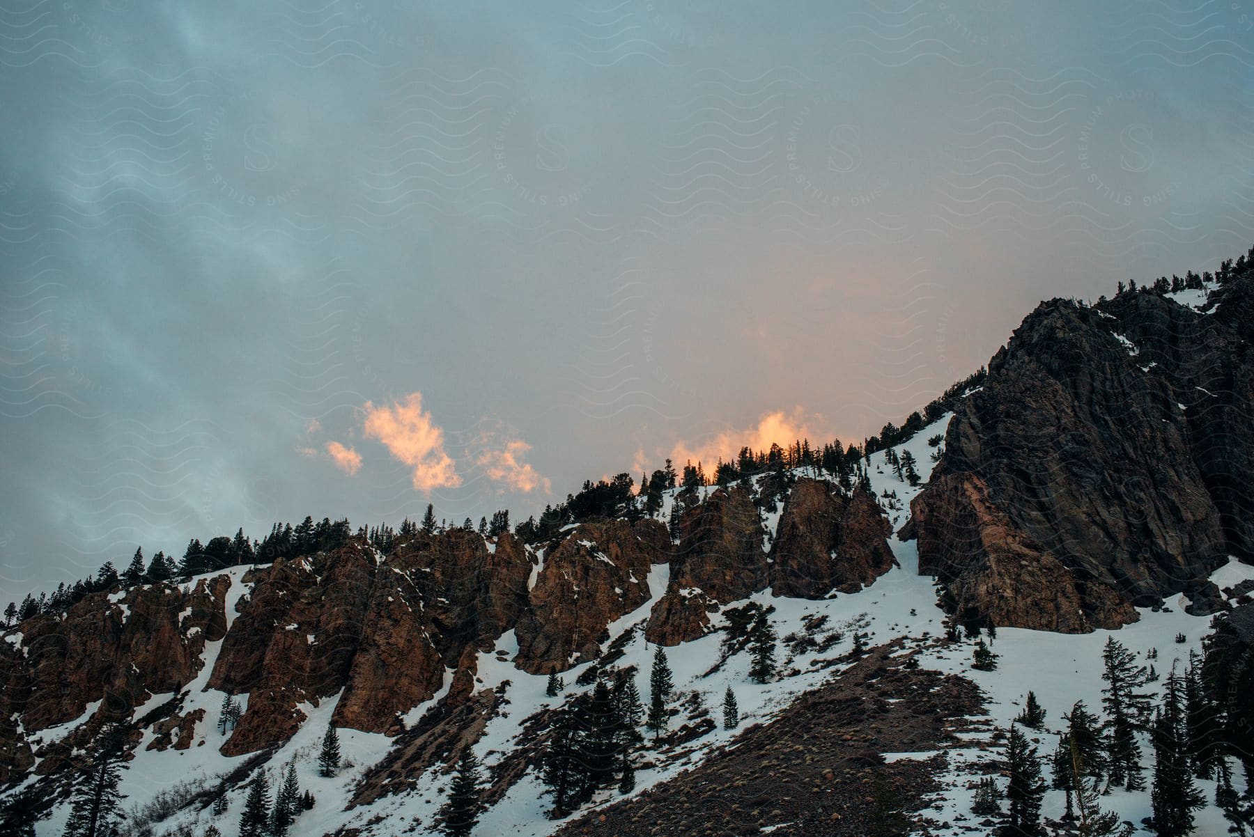 A rocky mountain is partly covered in snow under a cloudy sky at sunset.