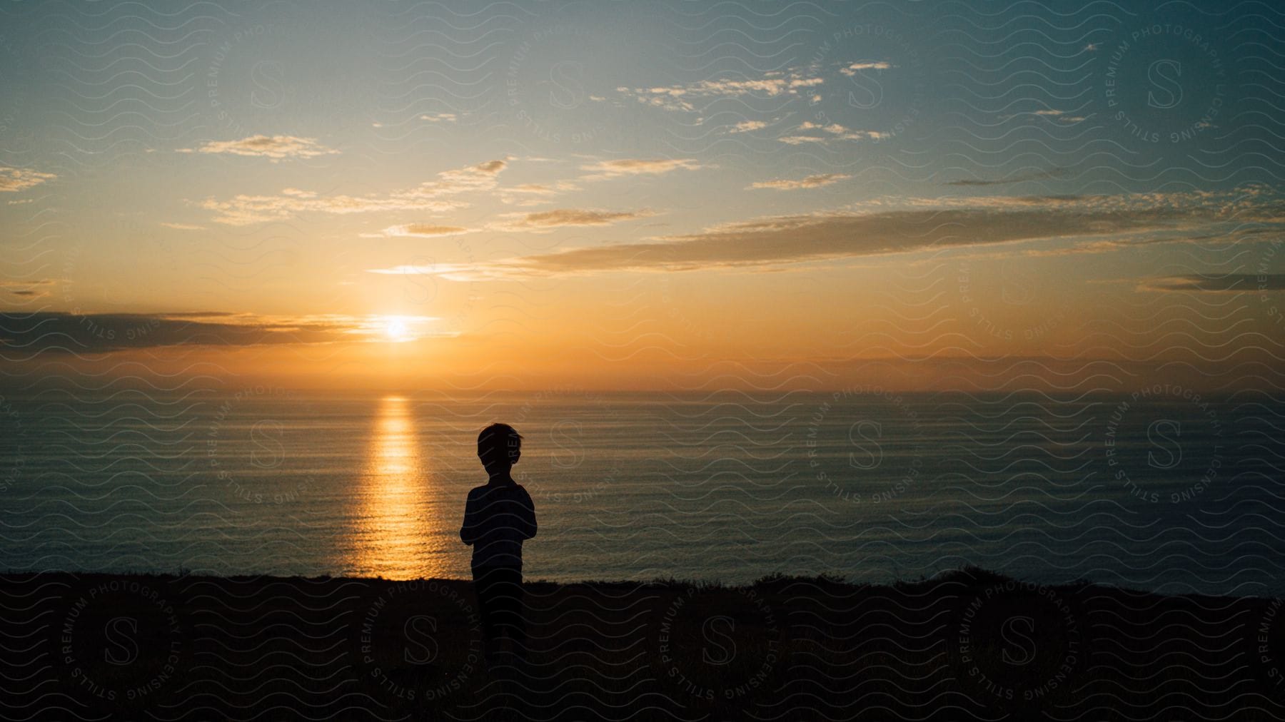 A young child is looking out at the water as the sun is setting.