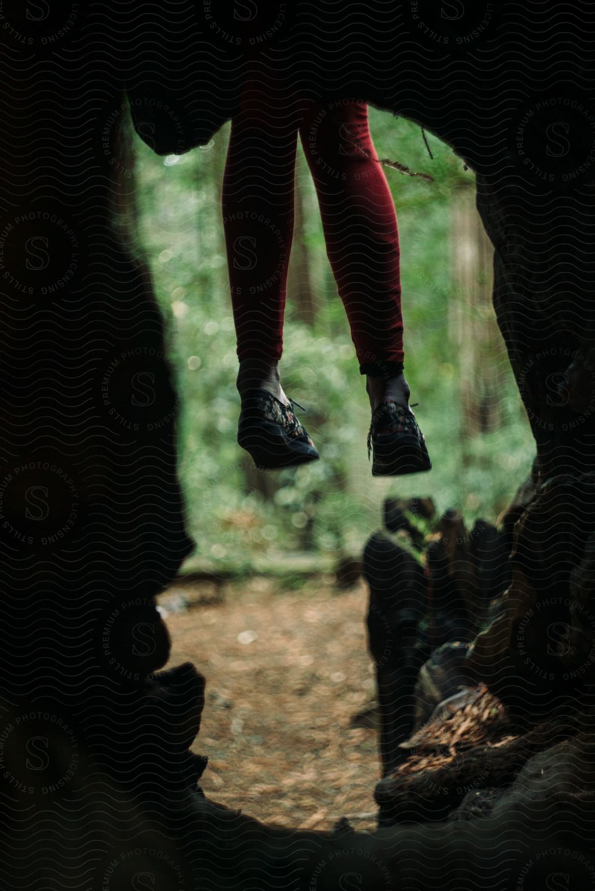 A girl, dressed in red pants and black shoes, sits on a cave in the forest