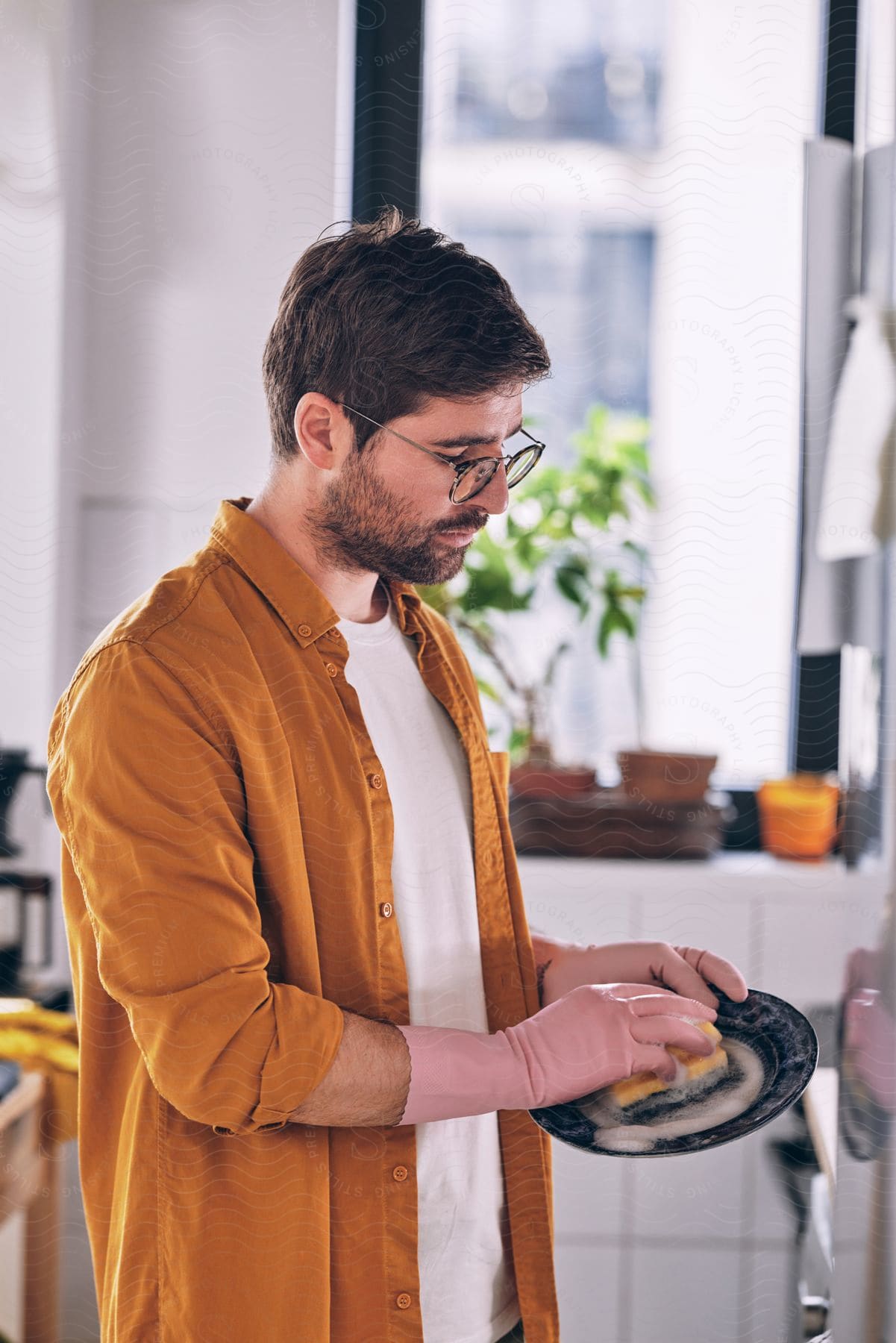 A bearded man with glasses, wearing a white top, pink gloves, and an orange shirt, is doing the dishes in the kitchen.