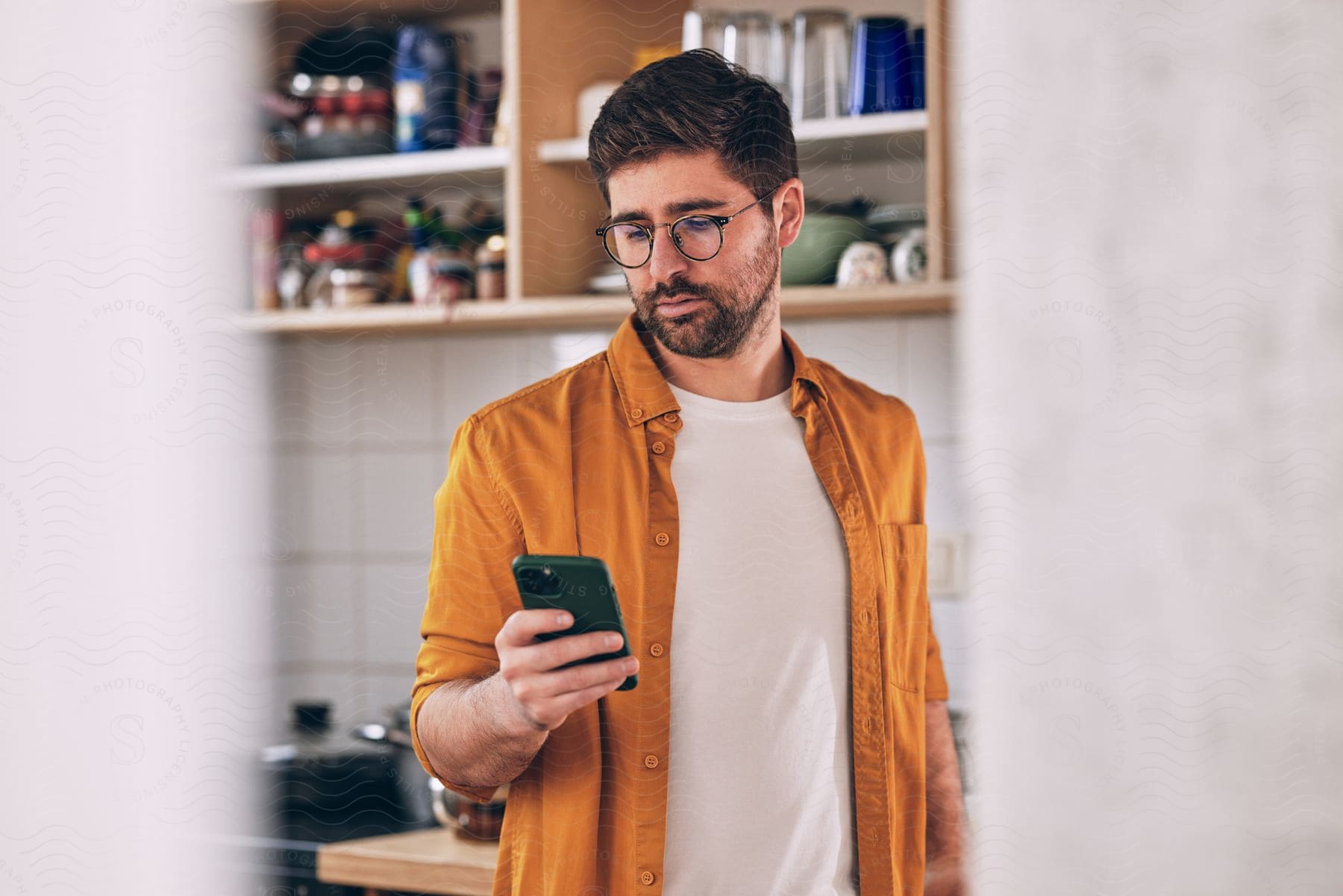 Stock photo of a man is looking at his cellphone as he stands in his kitchen.